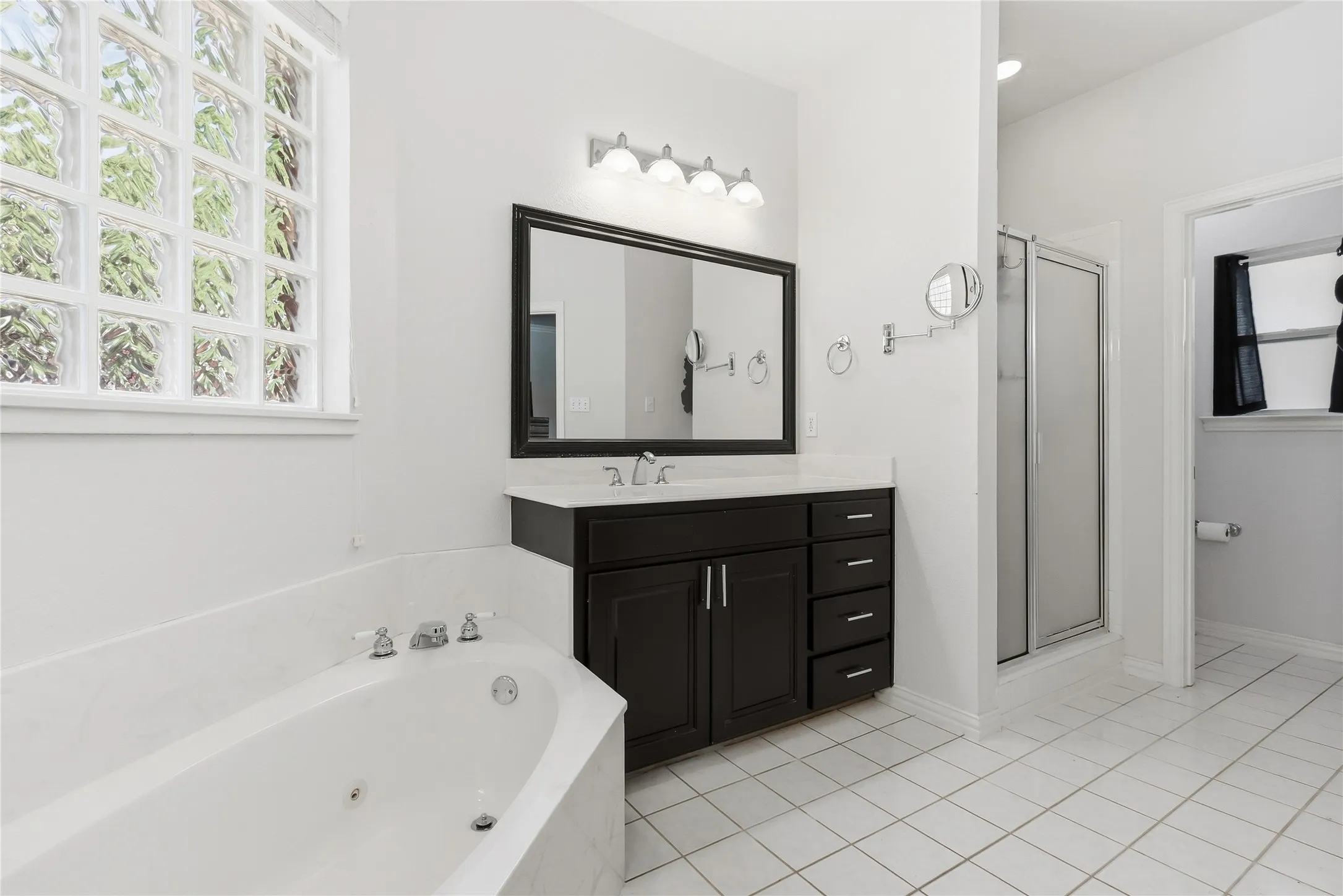 White tile flooring, corner jetted tub, glass block window, dark wood-finish vanity with countertop, and glass-enclosed shower