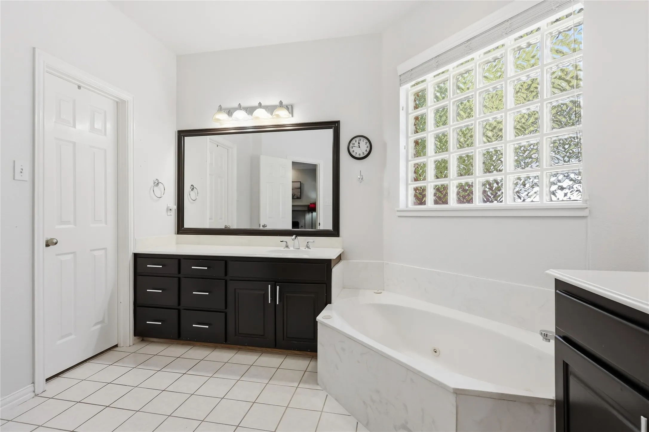 Expansive bathroom featuring a corner soaking tub, a wide vanity with a single basin, dark wood-finish cabinetry, a large framed mirror, and a privacy glass block window
