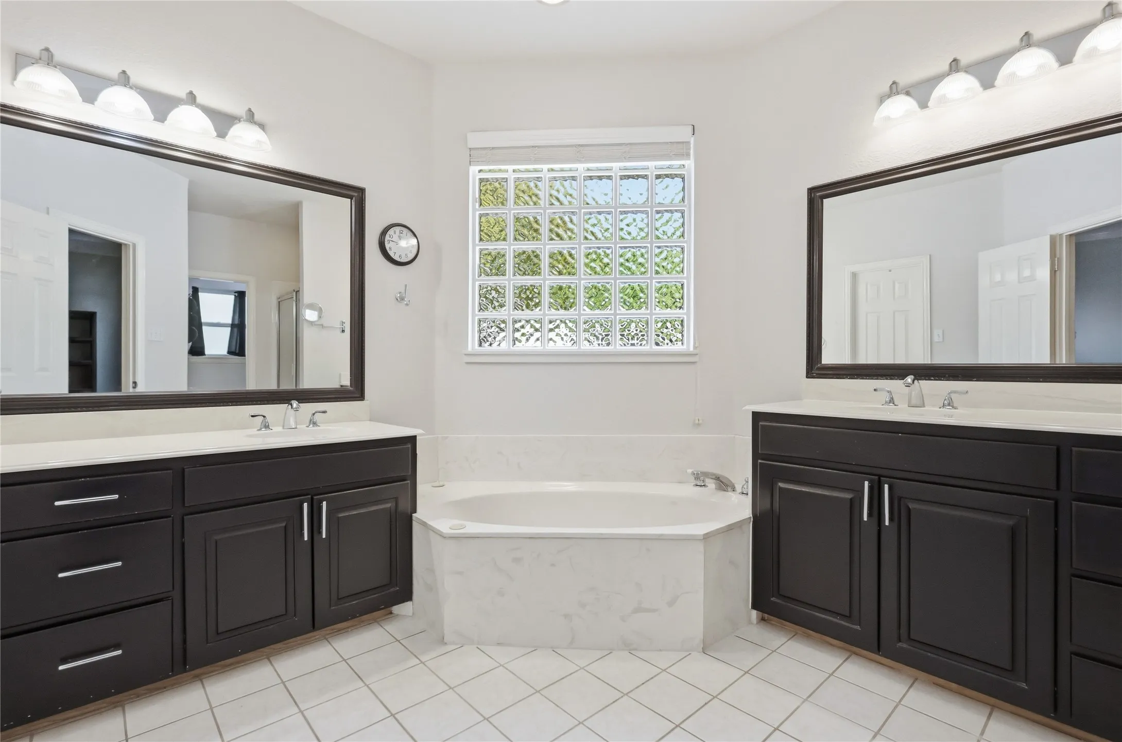 White tile flooring, dual dark wood-finish vanities with light countertops, large framed mirrors, and a built-in soaking tub with a tiled surround
