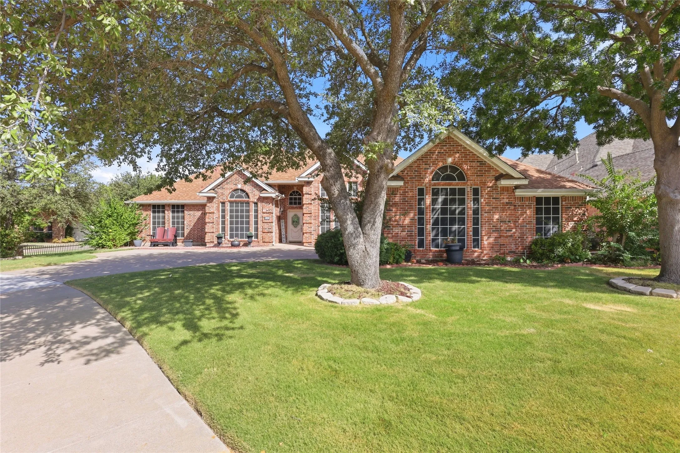 Red brick facade with arched window, established front yard, mature trees, and a concrete driveway