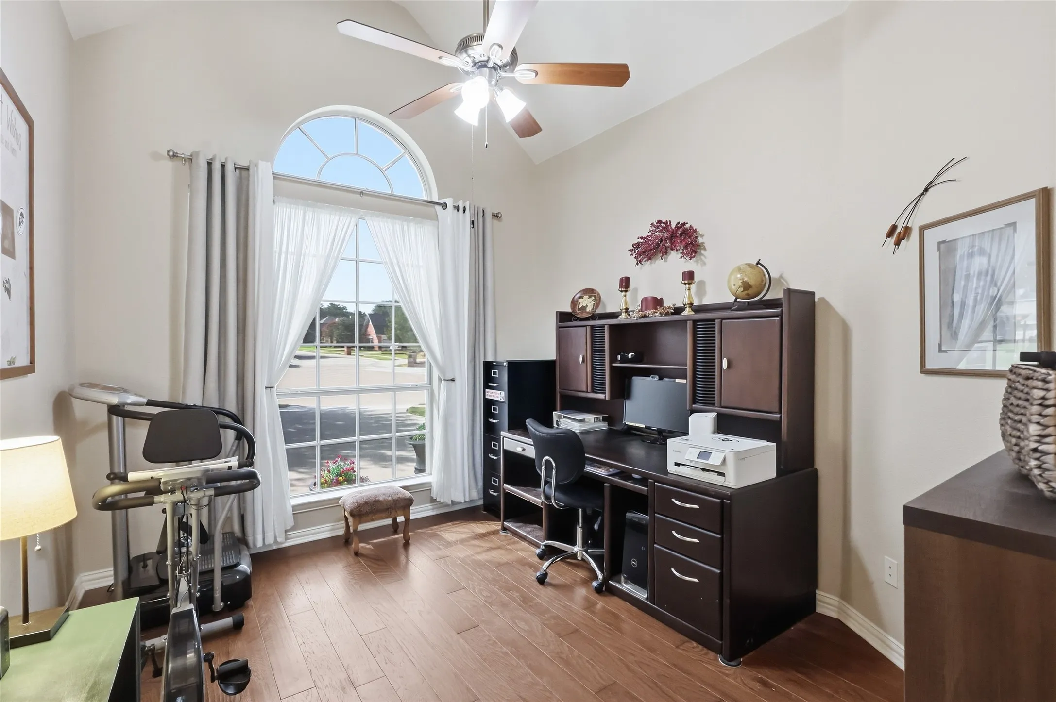 Arched transom window above a large picture window, vaulted ceiling, wood-finish flooring, and a ceiling fan with light fixture
