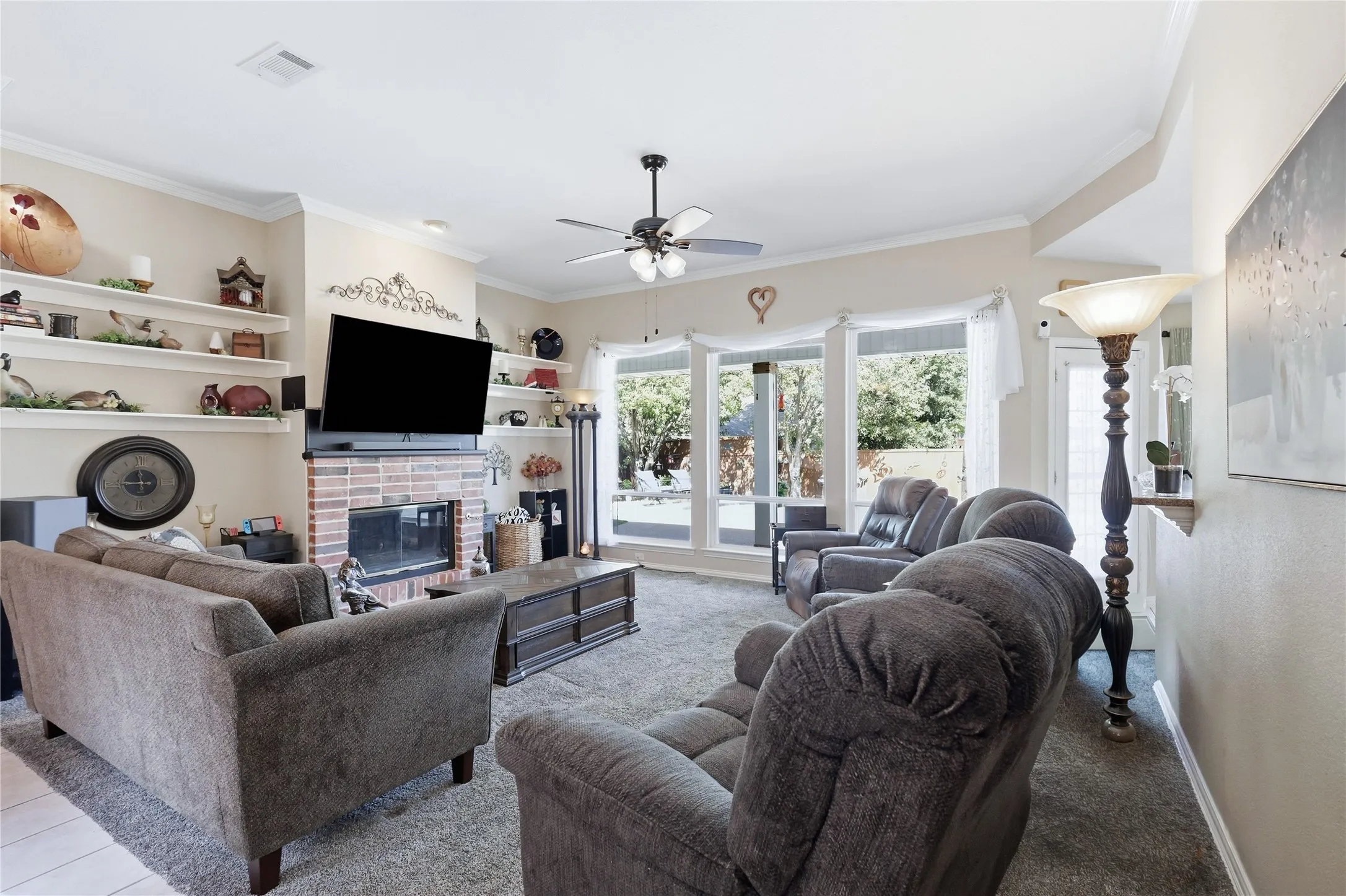Brick fireplace with a built-in television mount, integrated shelving, crown molding, a ceiling fan, and large windows