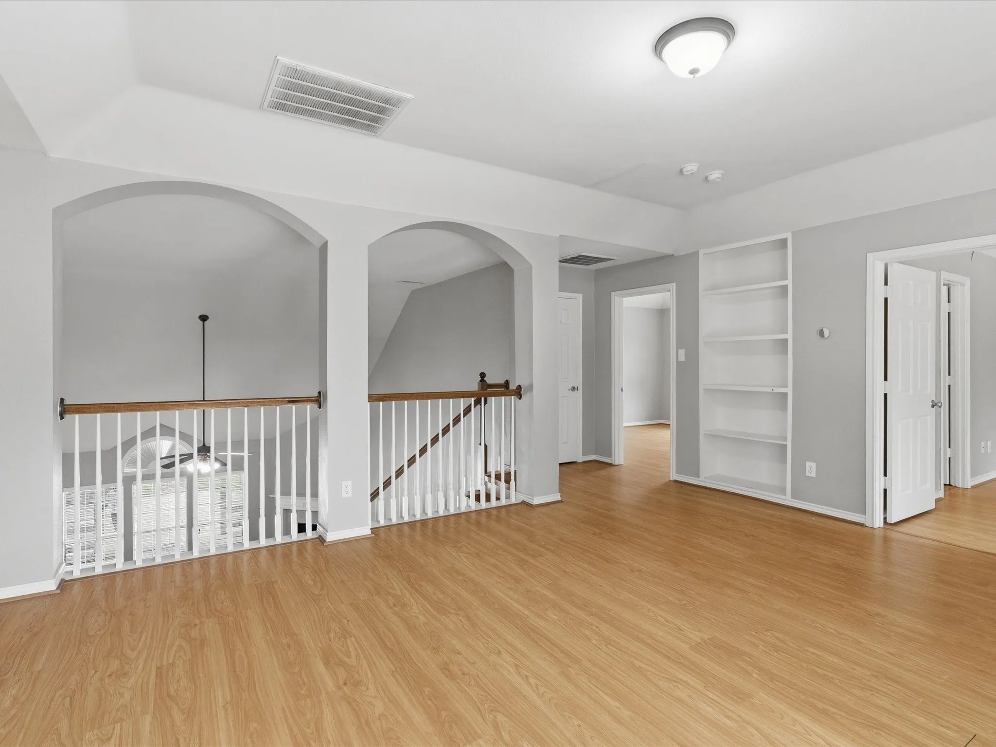 Wood-finish flooring throughout, featuring a built-in shelving unit, arched architectural elements, and a white spindle railing