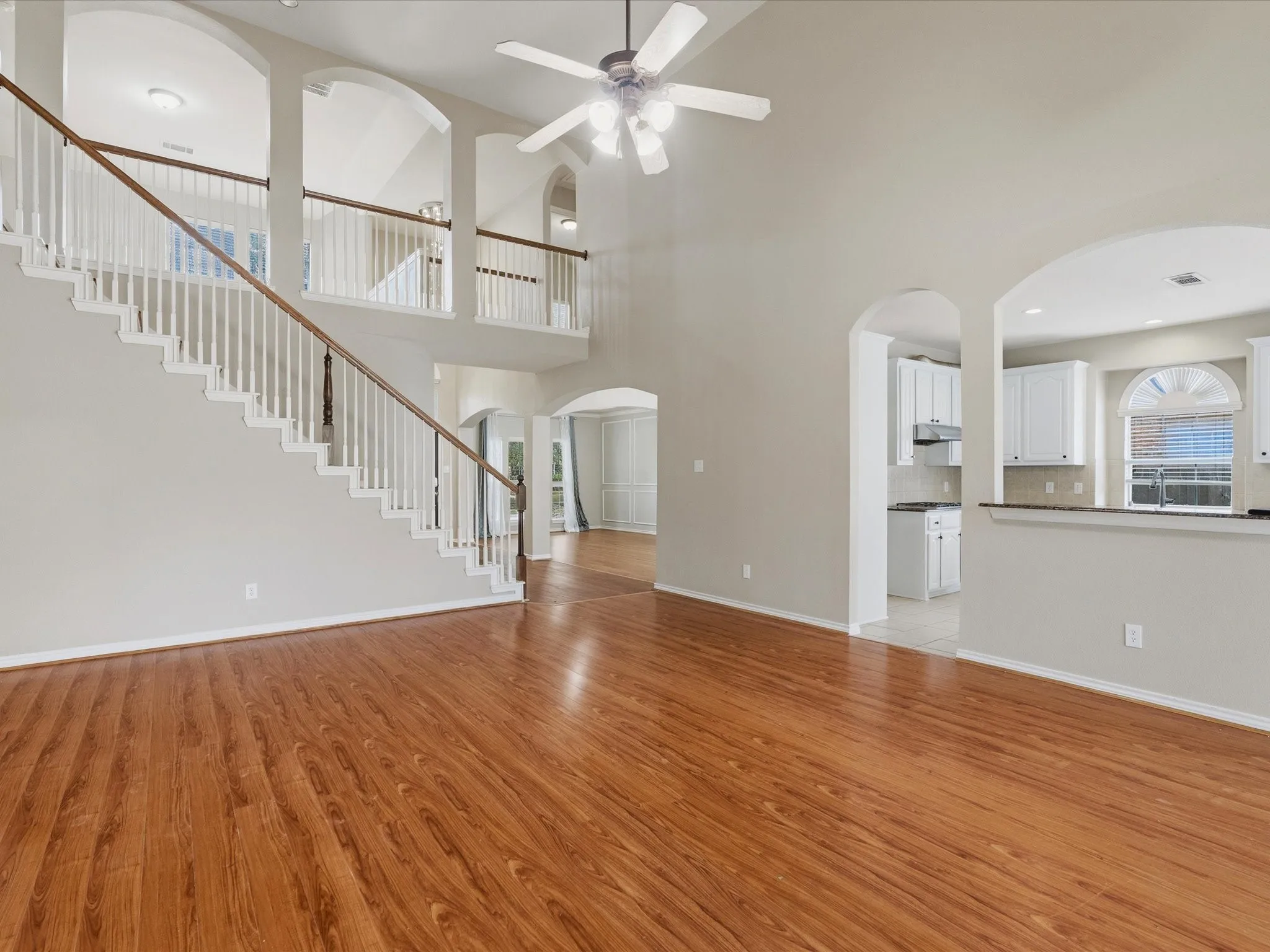 Two-story ceiling, wood-finish flooring, white spindle staircase with wood handrail, arched architectural details, kitchen with white cabinetry