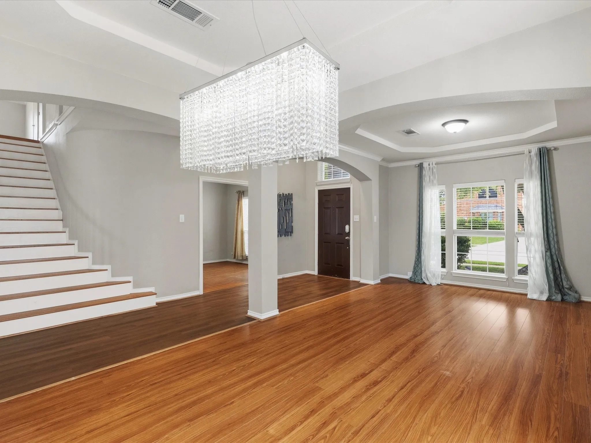 Wood-finish flooring throughout, tray ceiling with recessed lighting, prominent crystal chandelier, arched doorway, and a dark-paneled entry door
