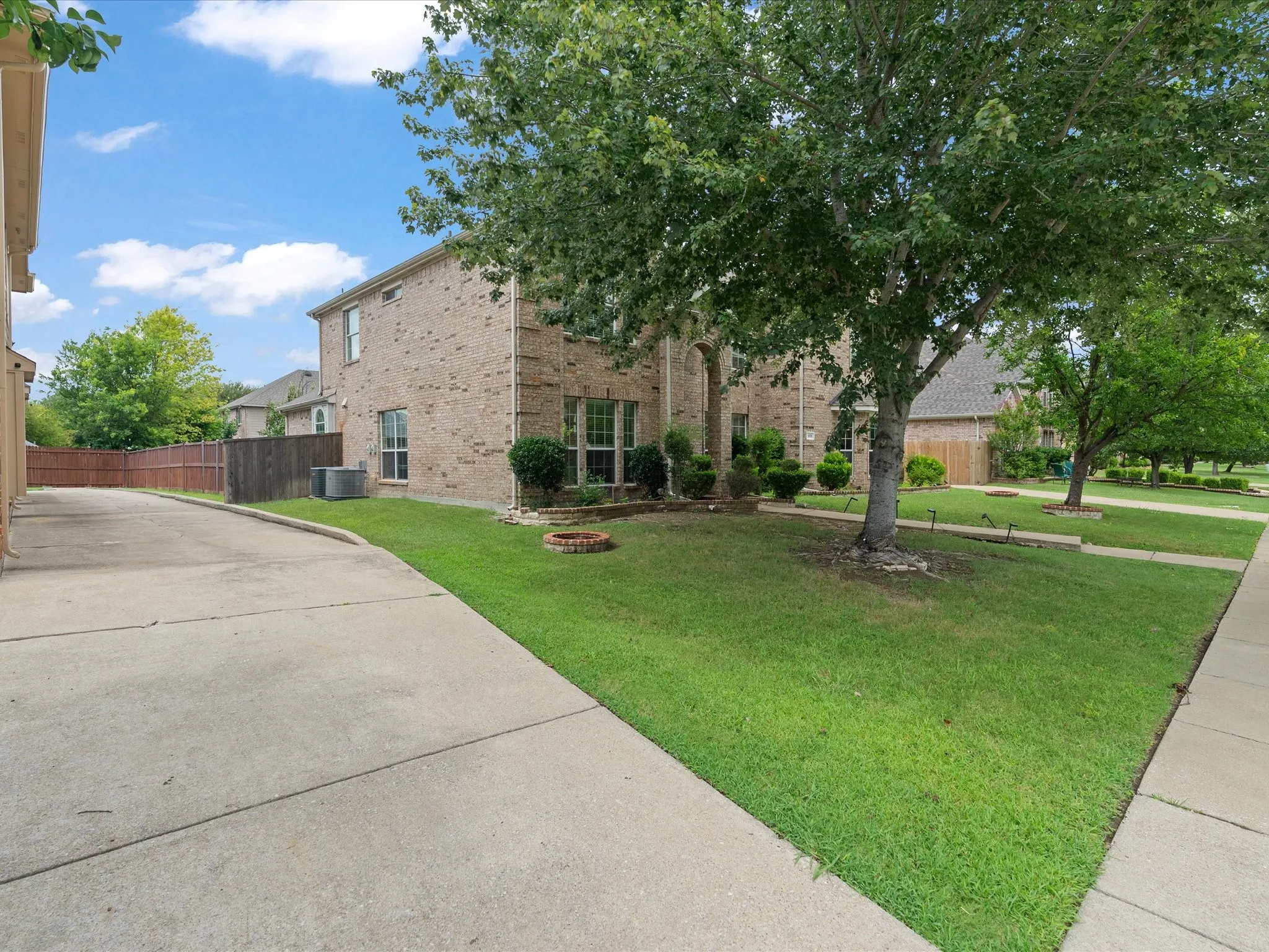 Brick-clad exterior with multiple windows, concrete driveway, lush green lawn, mature trees, and landscaped shrubbery