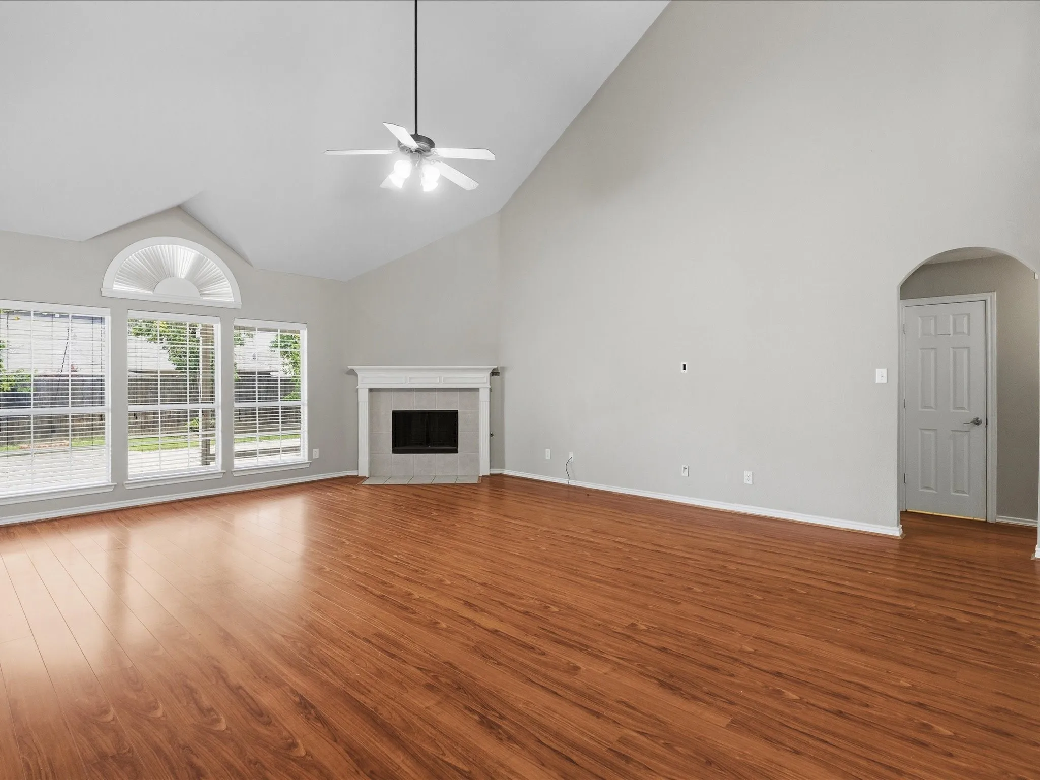 Vaulted ceiling with a ceiling fan, wood-finish flooring, multiple windows with blinds, a fireplace with a mantel, and an arched doorway