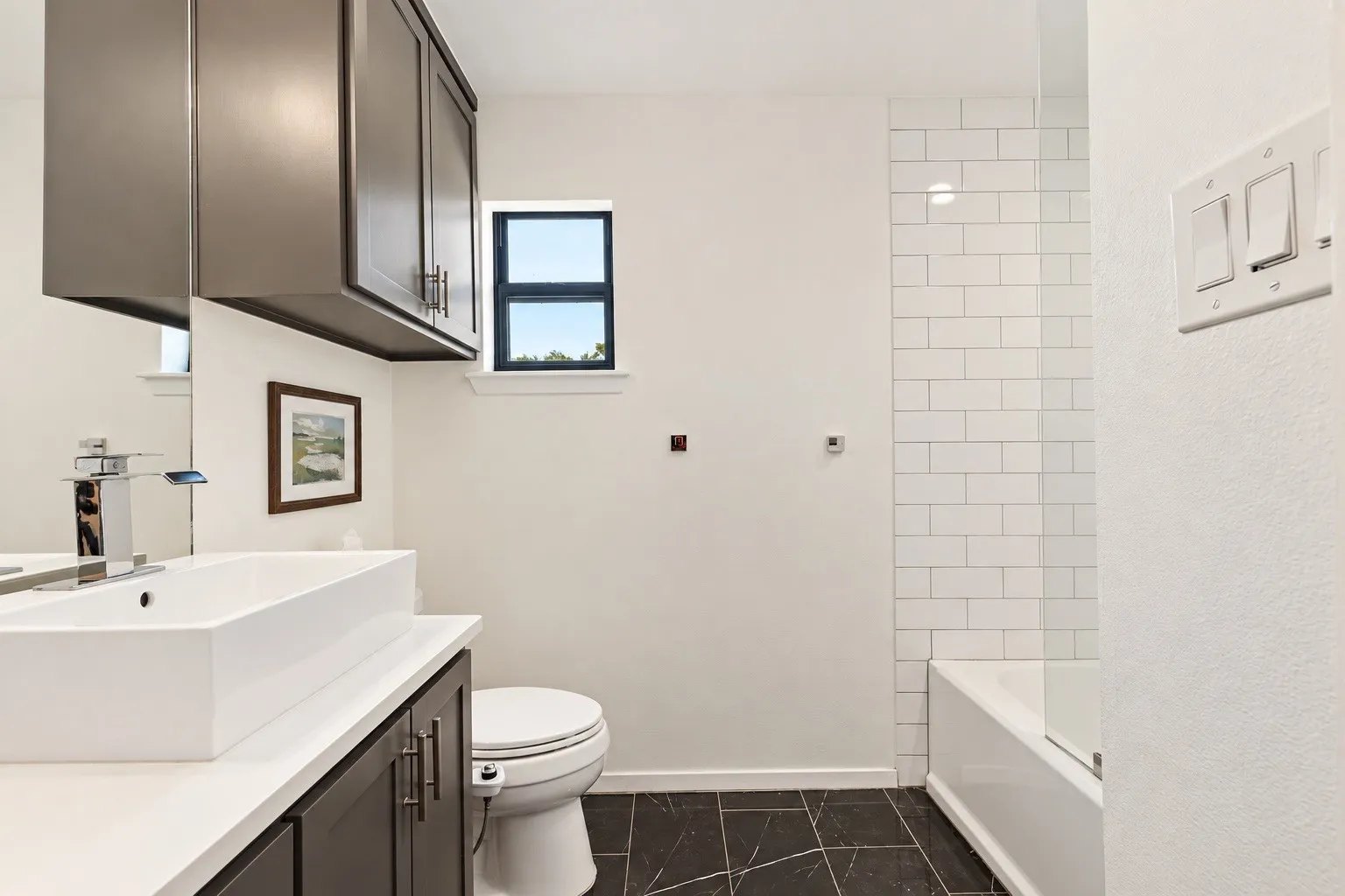 Vessel sink on a white countertop, dark wood-finish vanity with upper cabinetry, black tile flooring, and subway tile shower surround