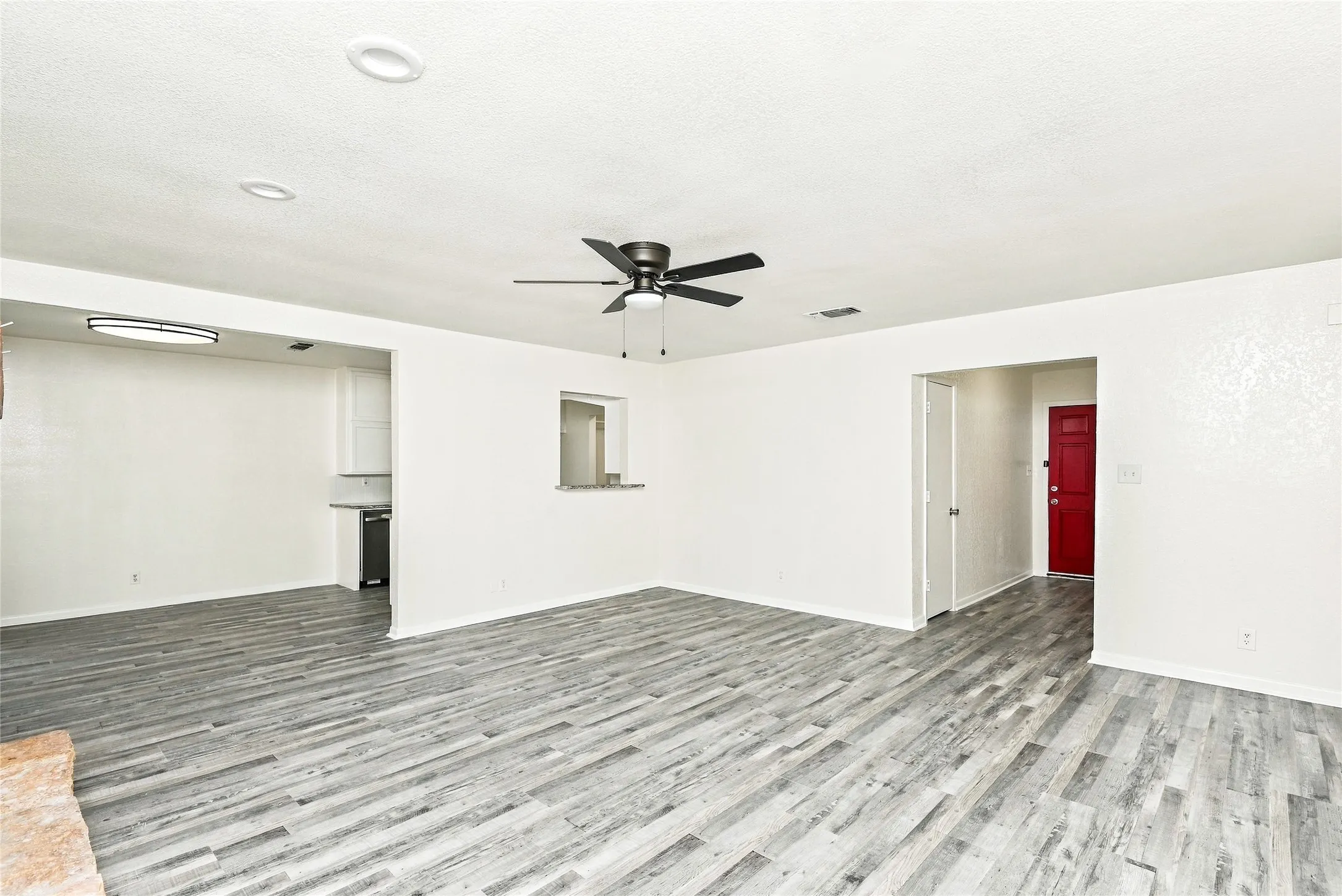 Wood-finish flooring throughout, recessed ceiling lighting, a dark-toned ceiling fan, a red-painted entry door, and a pass-through window to the kitchen