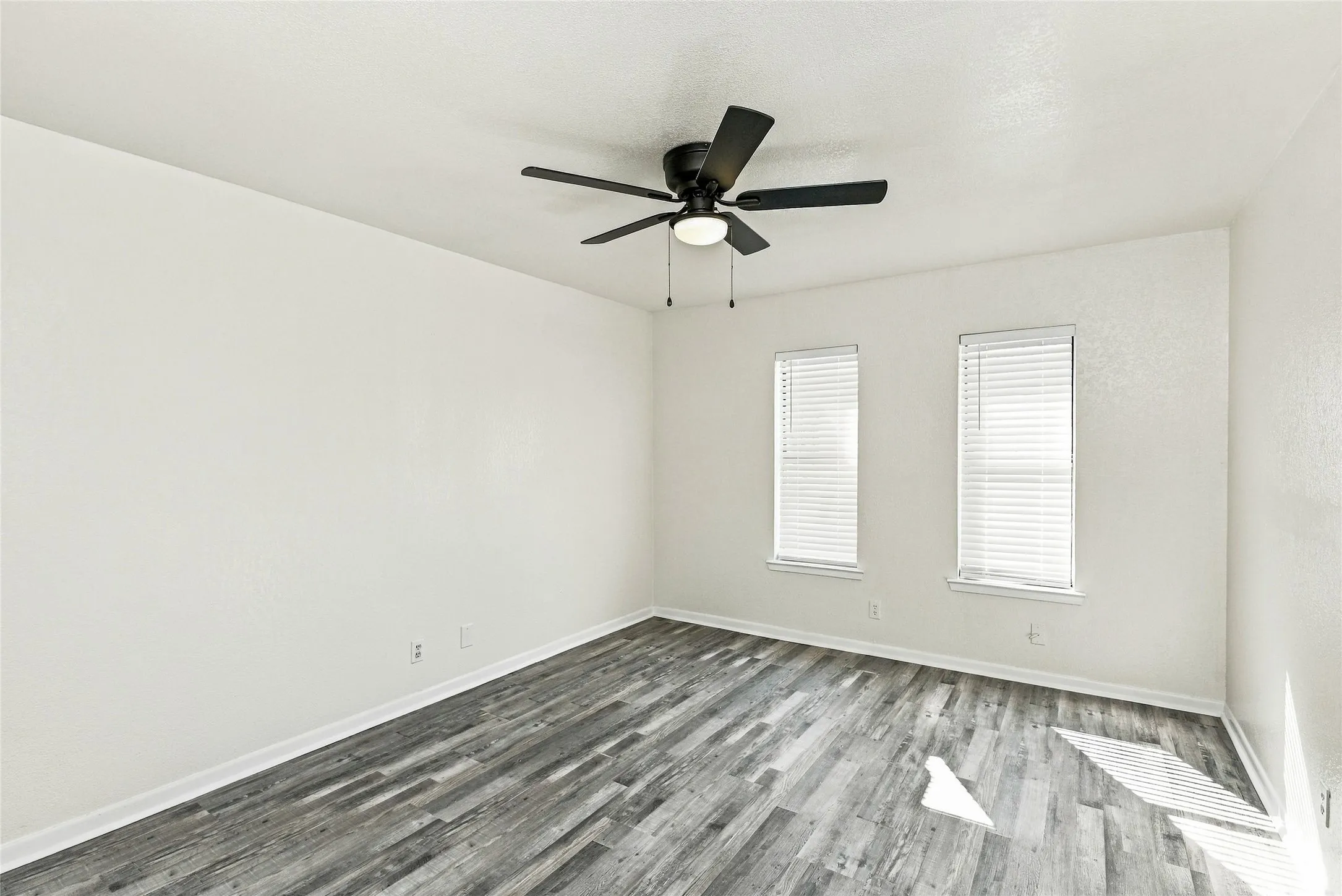 Wood-finish flooring, white walls, two windows with blinds, and a ceiling fan with integrated lighting