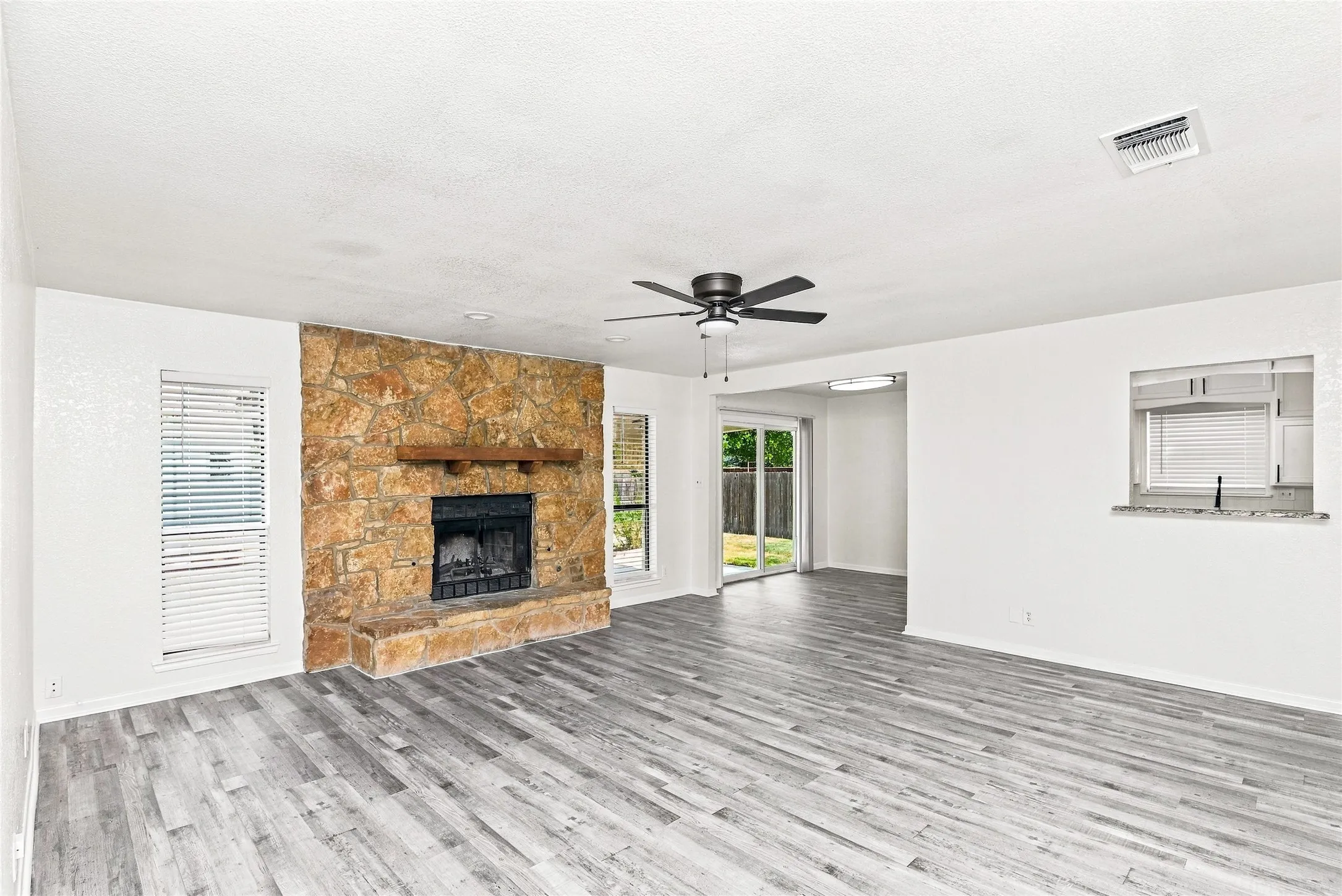 Stone-clad fireplace with wood mantel, wood-finish flooring, ceiling fan with integrated lighting, sliding glass door, and a pass-through opening
