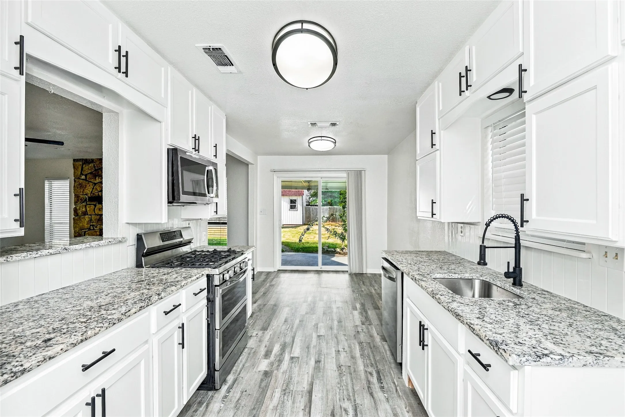 White shaker cabinetry with matte black hardware, stone-finish countertops, stainless steel appliances, wood-finish flooring, and sliding glass door access to the exterior