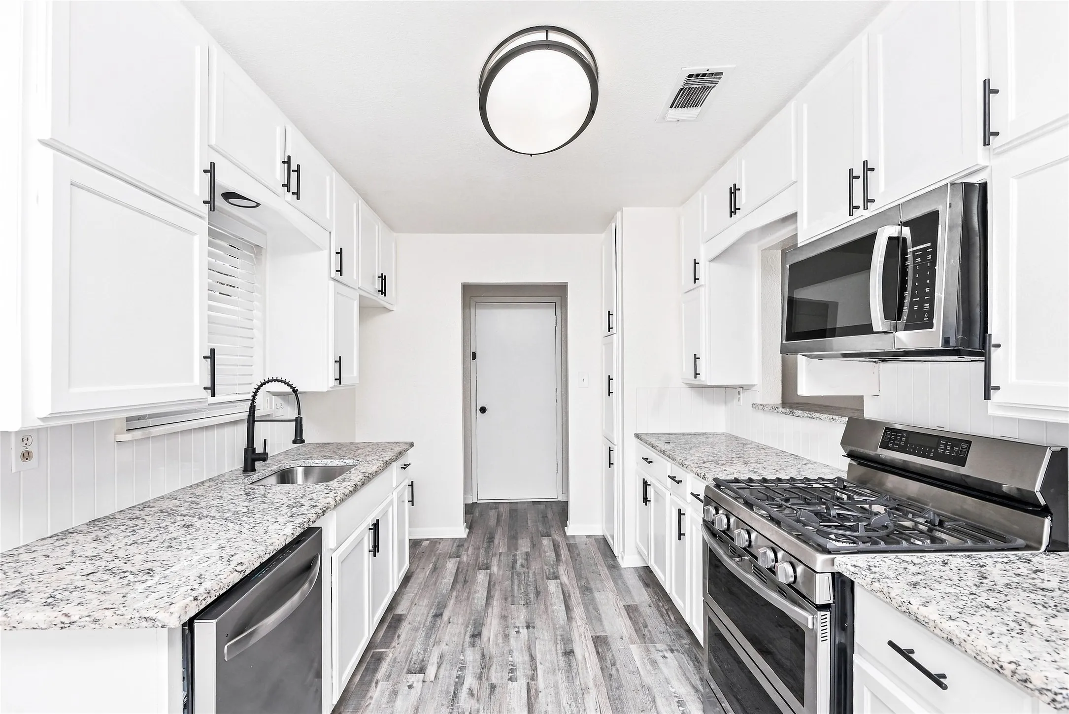 Galley kitchen featuring white cabinetry, light-toned countertops, stainless steel appliances, a matte black faucet, and wood-finish flooring