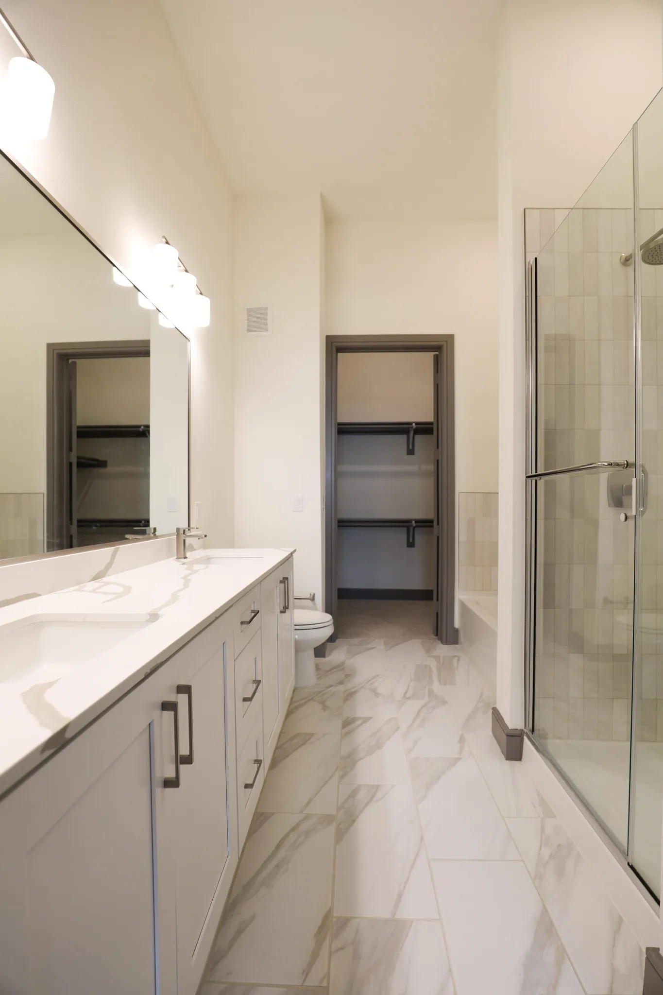 Dual vanity with integrated sinks, large mirrored wall, tile flooring, glass-enclosed shower, and built-in closet shelving