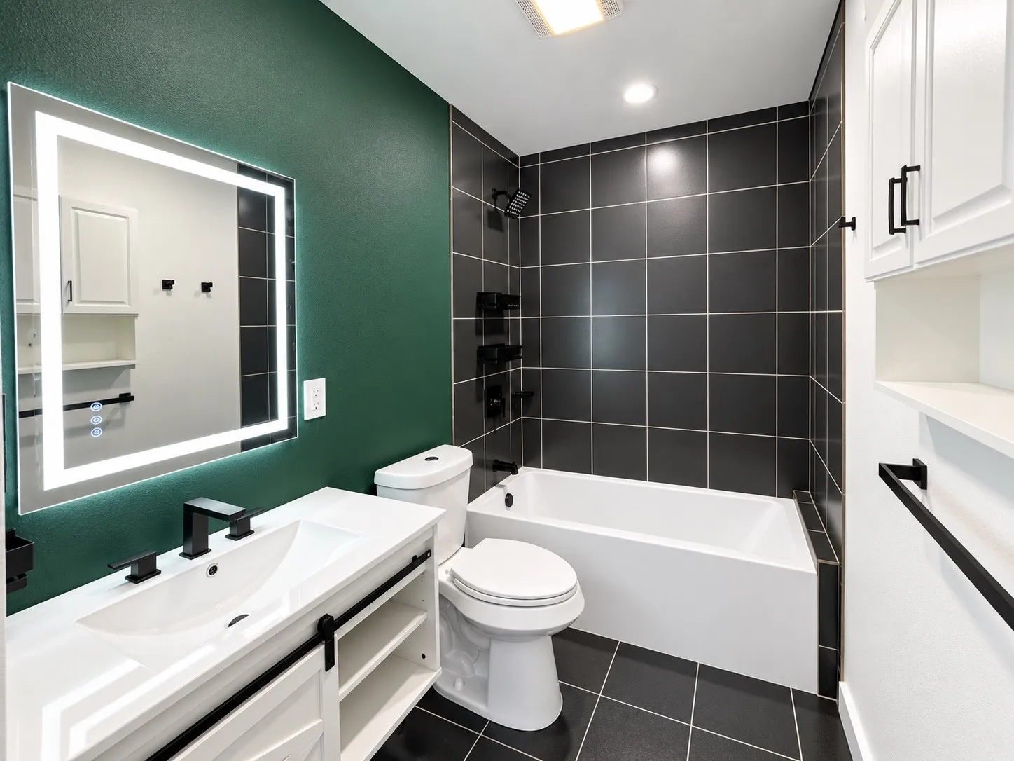 Illuminated vanity mirror, rectangular undermount sink, white vanity with barn door style cabinetry, black tile shower surround, and a white bathtub