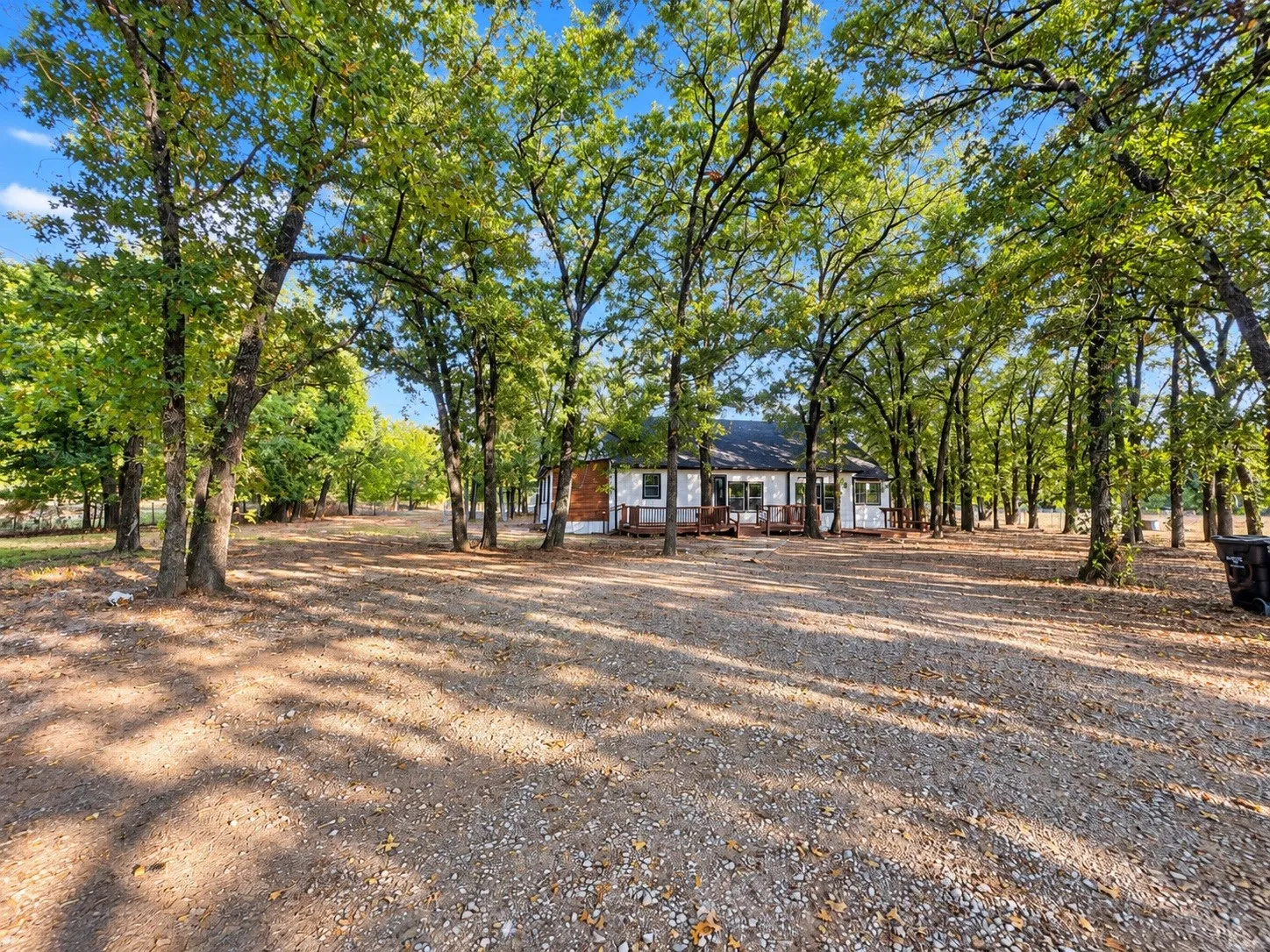 Expansive gravel drive leading to a residence featuring a white exterior with dark trim, multiple windows, and a dark shingle roof, all surrounded by mature trees