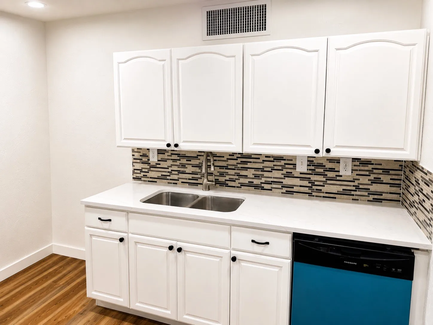 Kitchenette featuring white cabinetry with black hardware, a mosaic tile backsplash, double basin stainless steel sink, and light-toned countertops