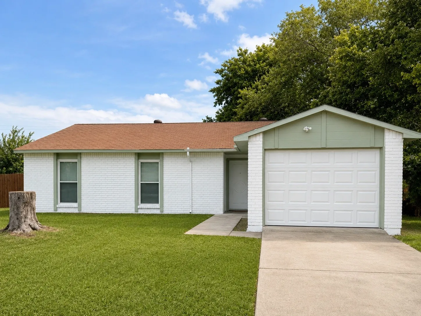 White brick exterior with a brown shingle roof, featuring a single-car garage with a panel door, a paved driveway, and a concrete walkway