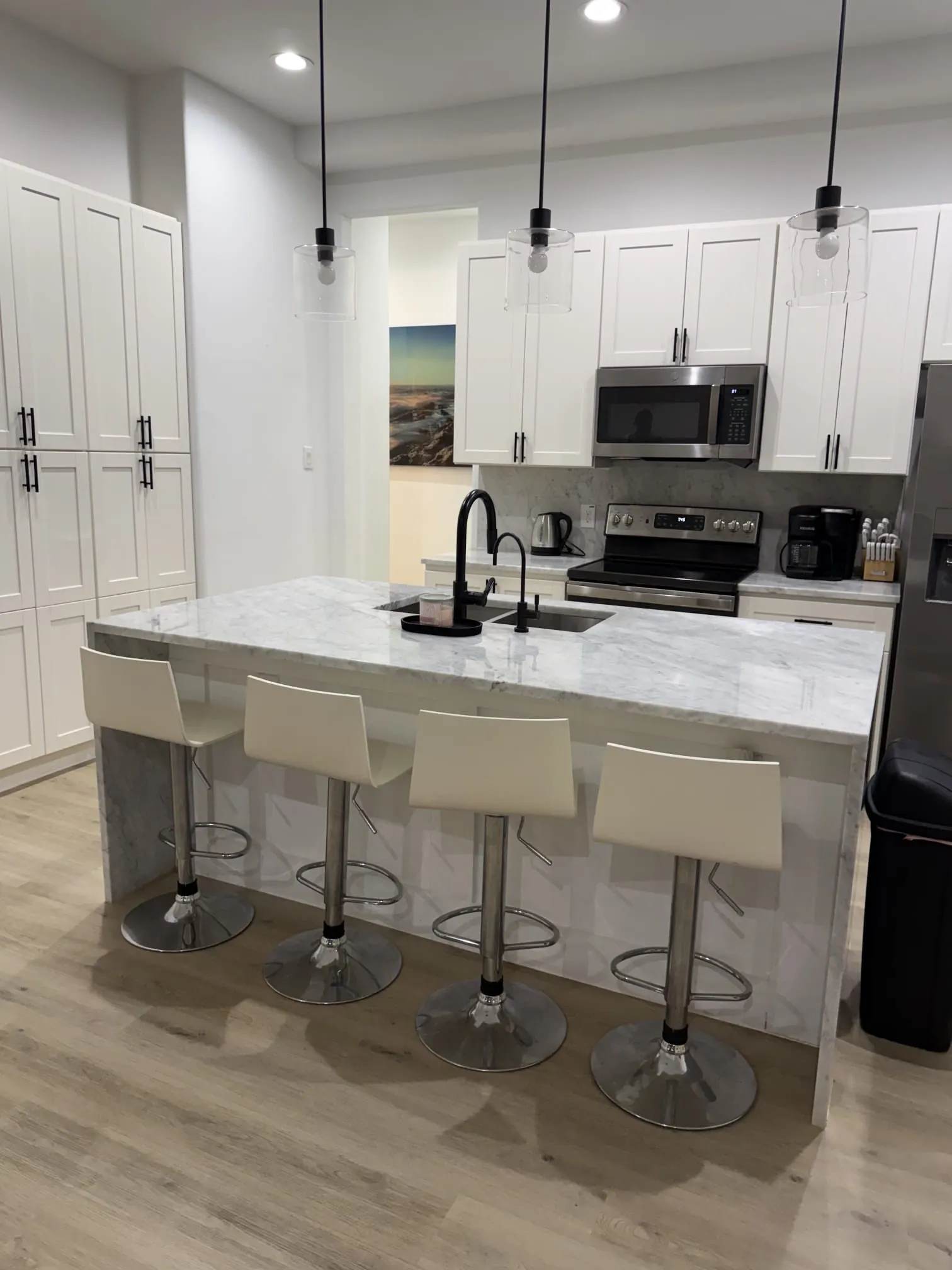 Kitchen featuring white shaker-style cabinetry, a waterfall island with a light-toned countertop, stainless steel appliances, and a black matte faucet