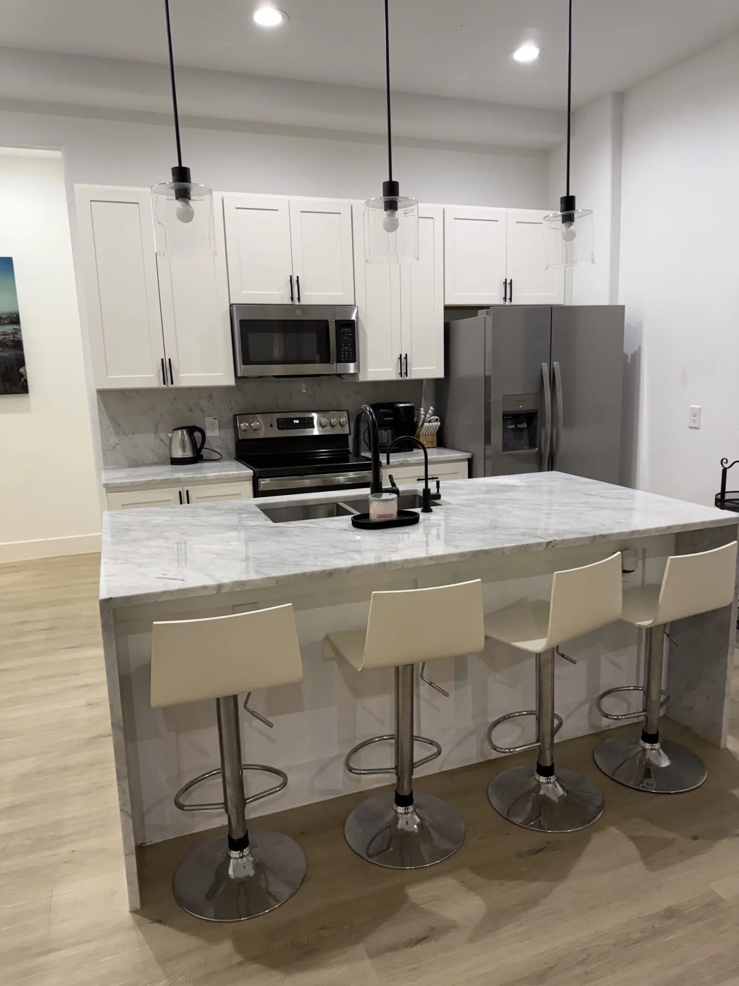Kitchen featuring a waterfall-edge island with integrated sink, stainless steel appliances, white cabinetry with dark hardware, and wood-finish flooring