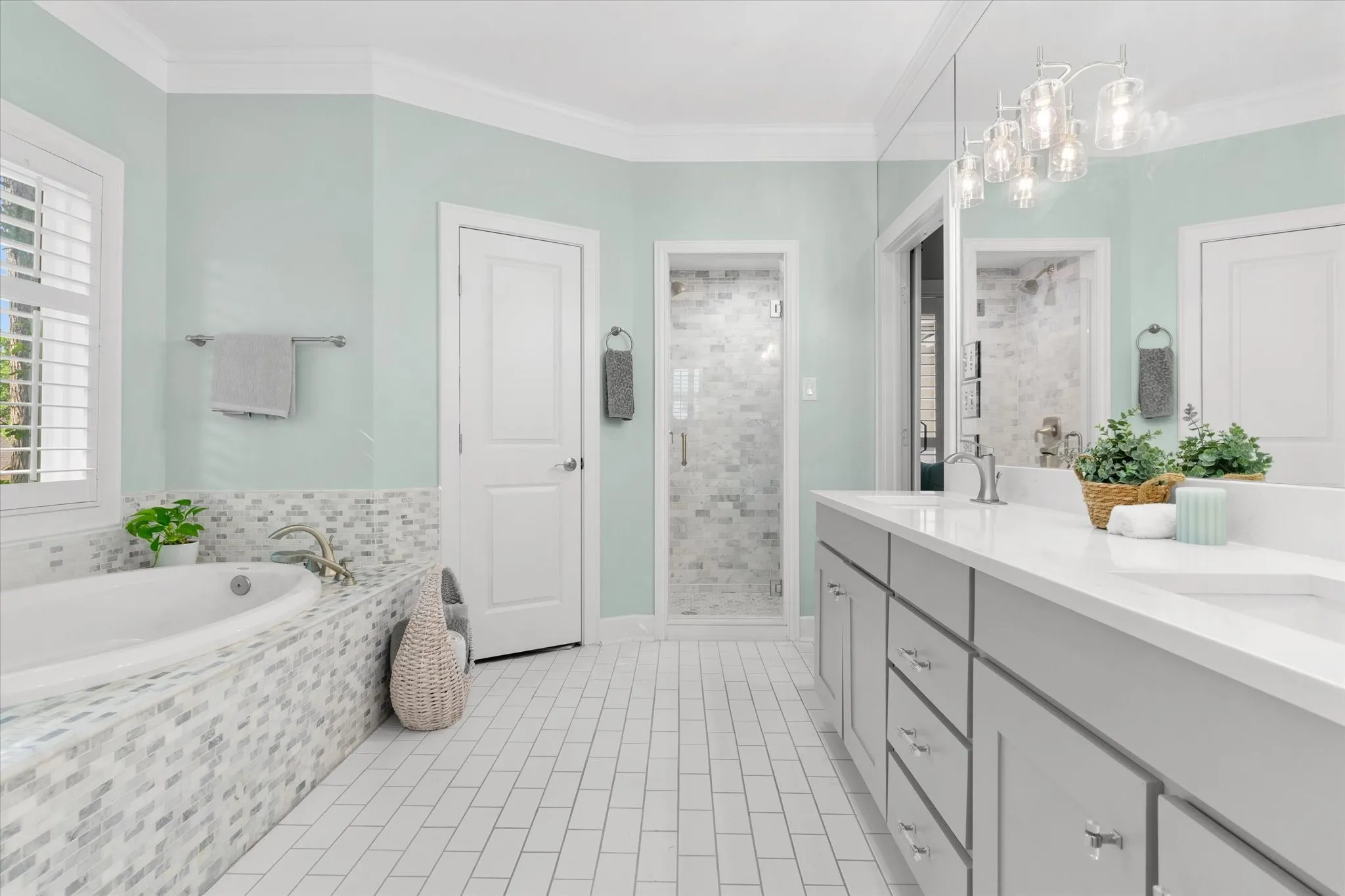 Large vanity with dual sinks, a built-in bathtub with mosaic tile surround, a glass-enclosed shower, wood shutters, and white subway tile flooring