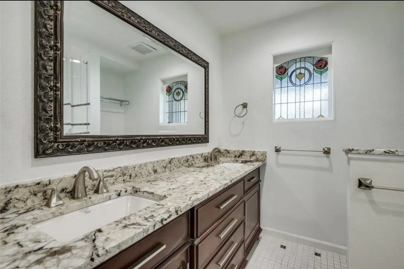 Dual undermount sinks, stone-finish countertop, dark wood-finish cabinetry, decorative stained-glass window, and white square tile flooring
