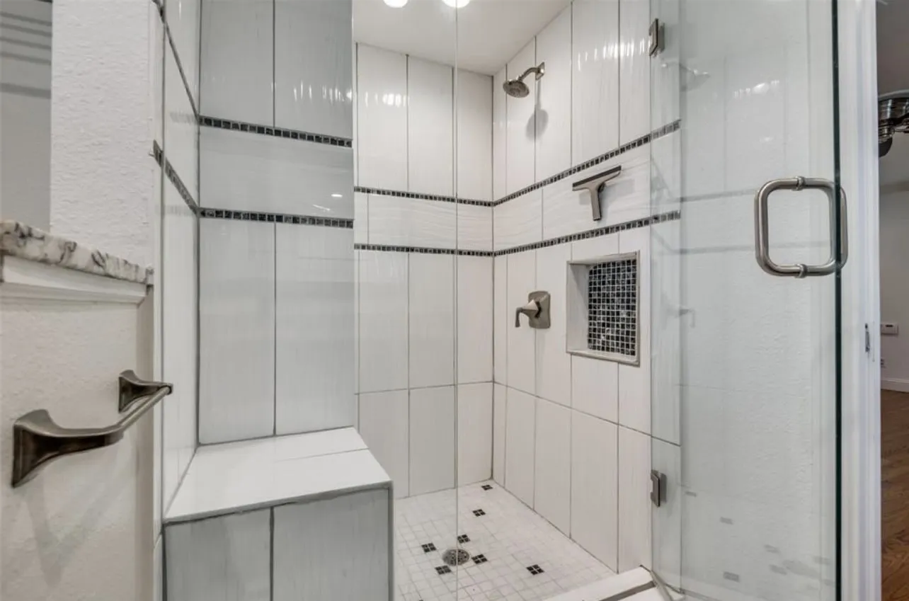 Glass-enclosed shower with white tiled walls, mosaic accent band, built-in bench, recessed shelf with mosaic tile, and brushed nickel fixtures