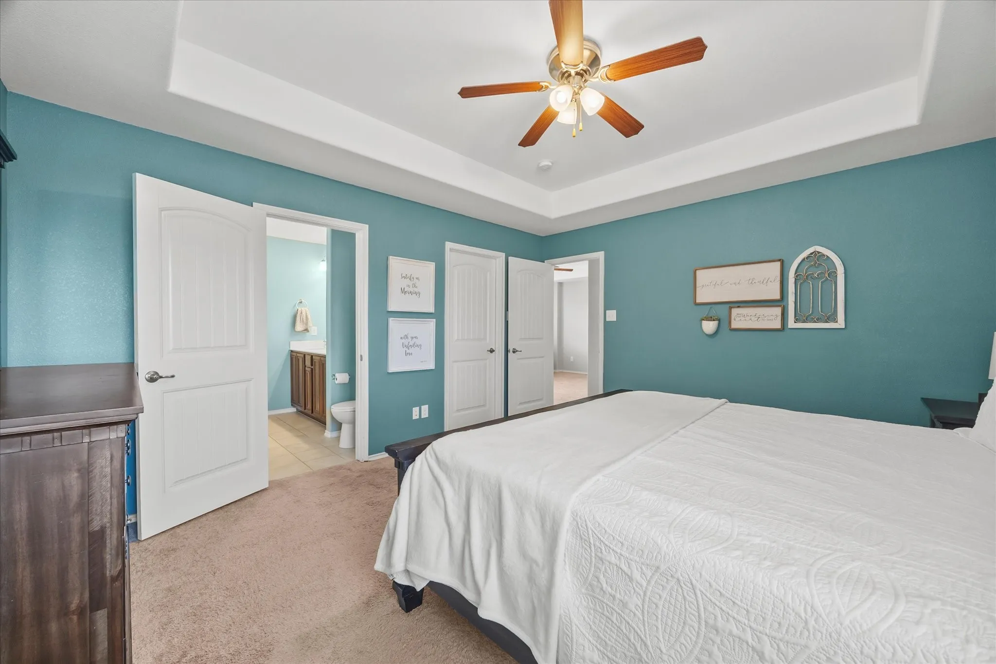 Tray ceiling, ceiling fan with light fixture, carpeted flooring, wood-finish doors, and an attached bathroom with a single vanity