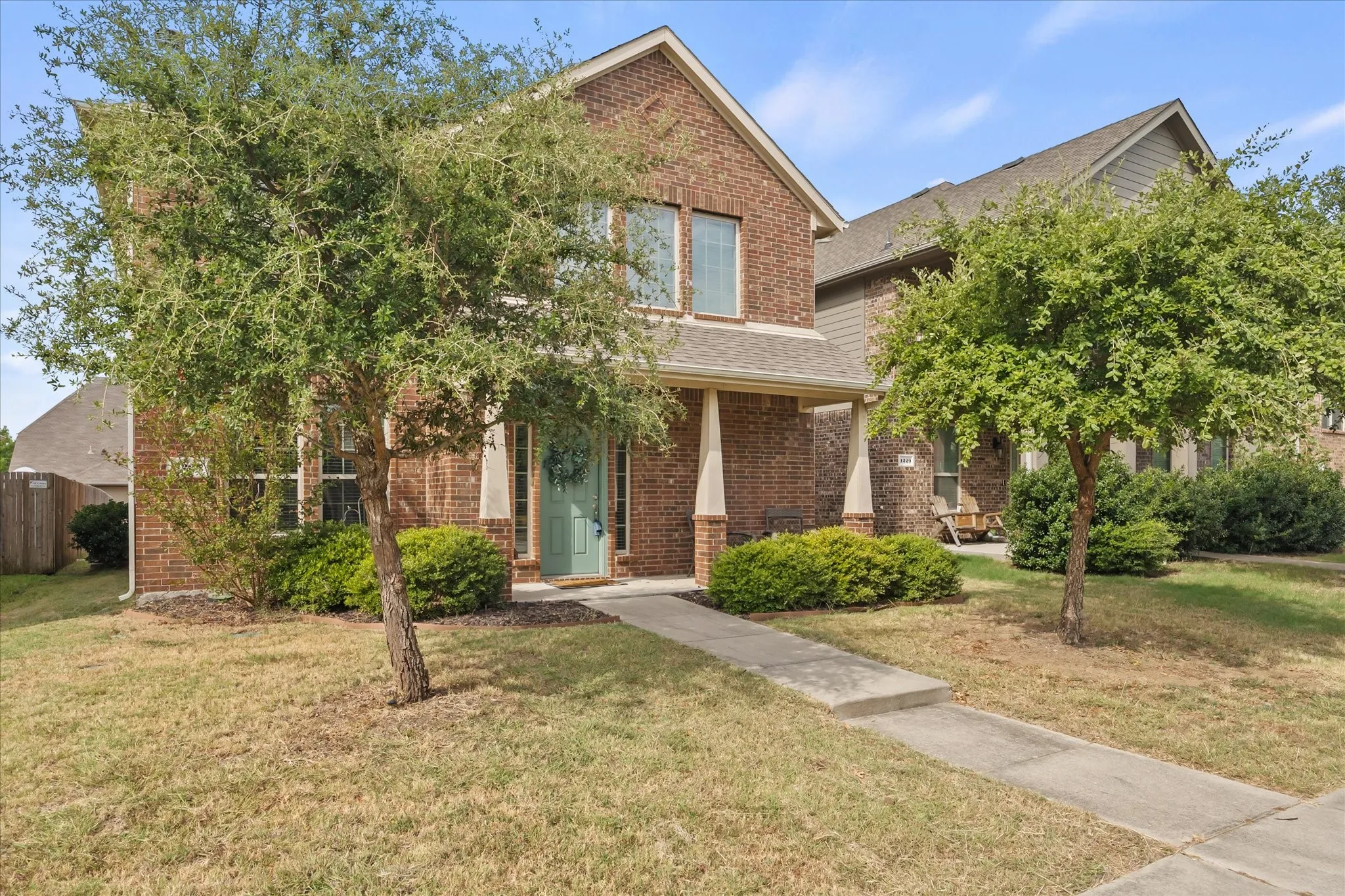 Brick exterior facade with a covered entry, established landscaping, a concrete walkway, and a partial privacy fence