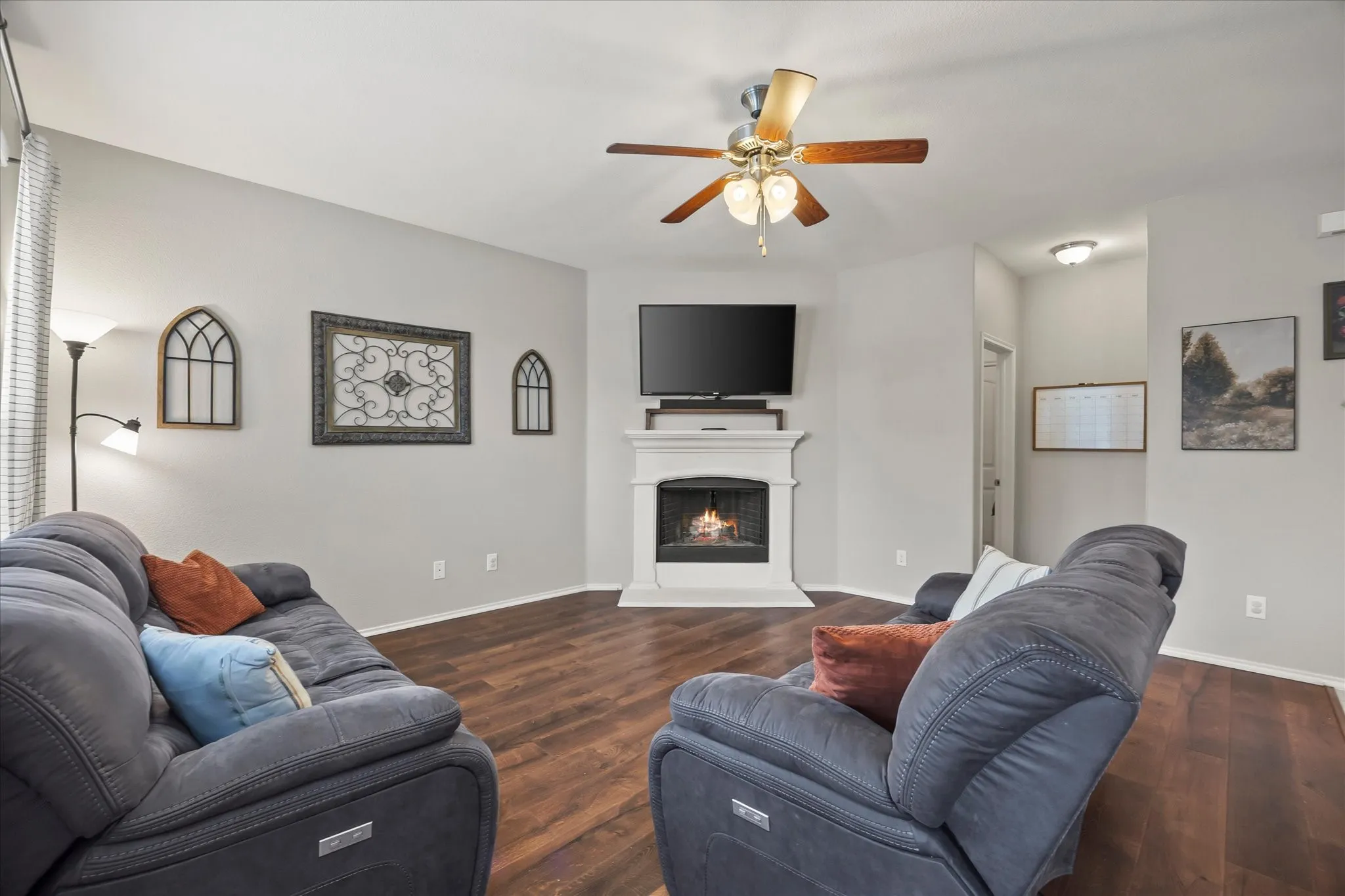 Wood-finish flooring, central fireplace with white mantel, ceiling fan with integrated lighting, recessed wall niche, and light gray wall paint throughout