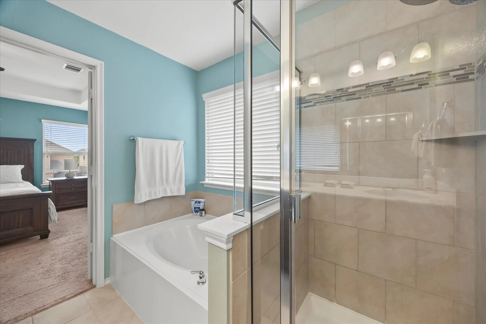 Built-in soaking tub, glass-enclosed shower with tiled walls, decorative tile shower accent, and a row of four vanity lights