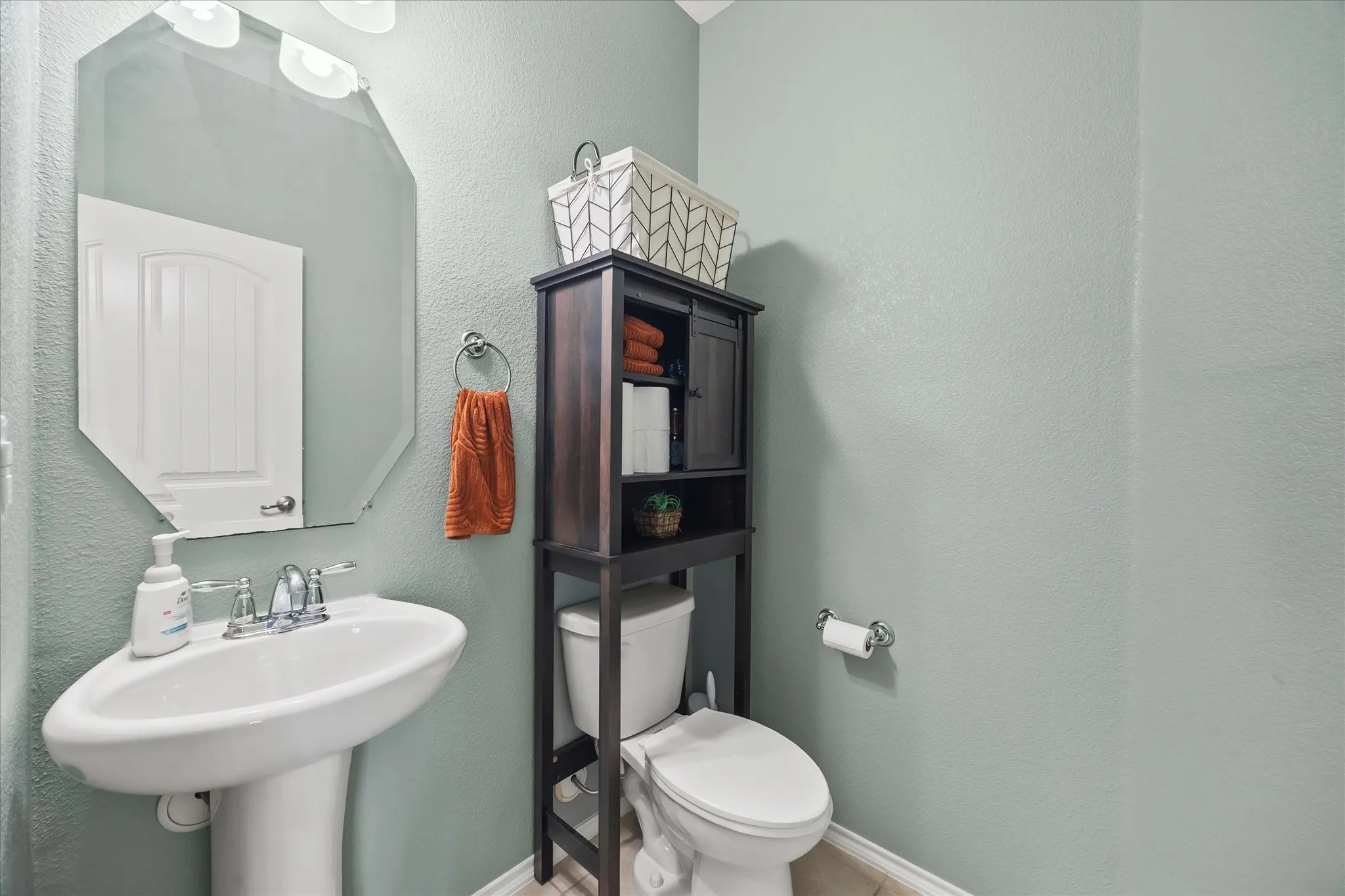 Pedestal sink with chrome faucet, octagonal wall mirror, and built-in over-the-toilet storage cabinet