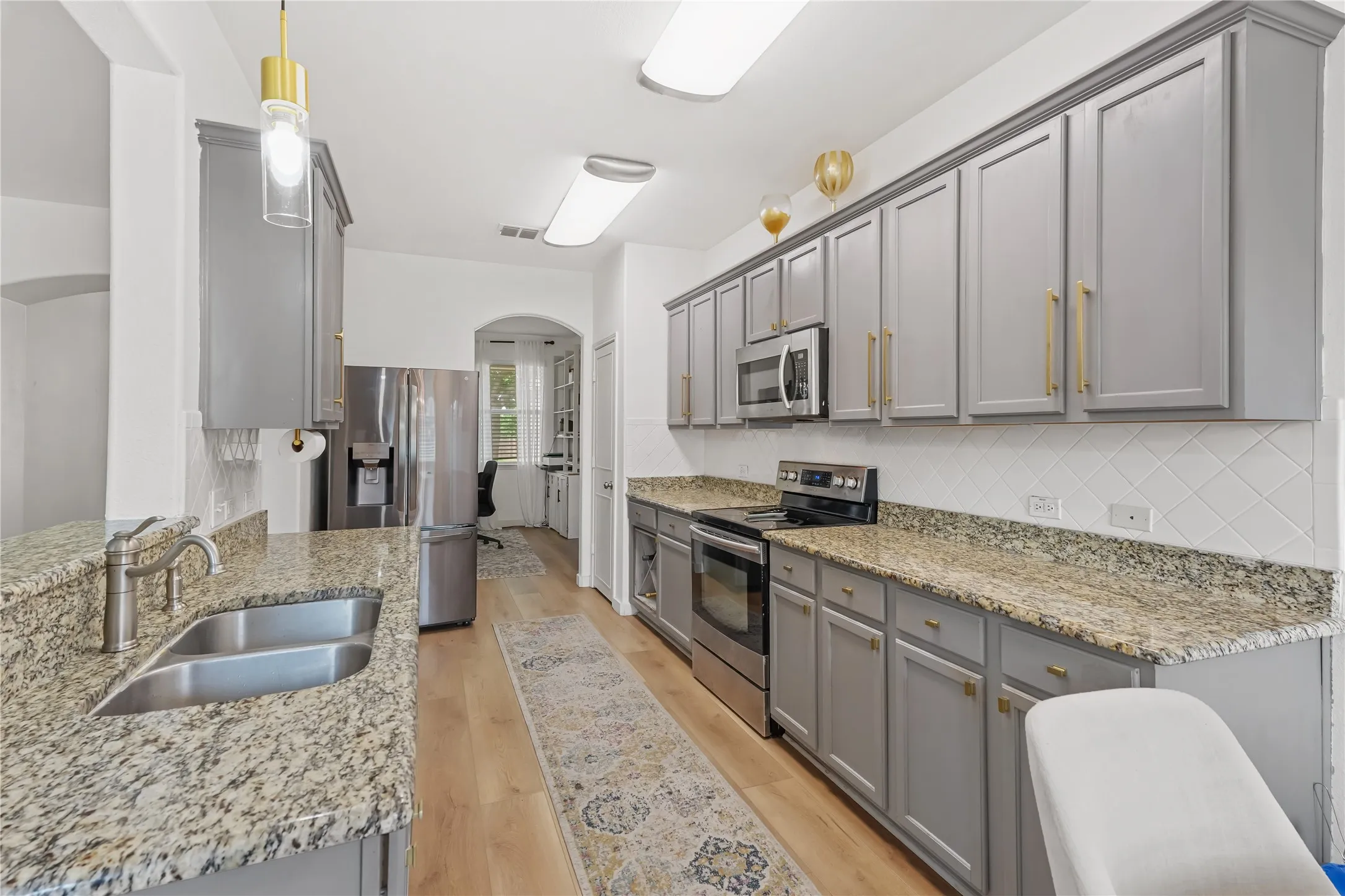 Stainless steel appliances, light gray cabinetry, light-toned wood-finish flooring, stone countertops, and white tile backsplash