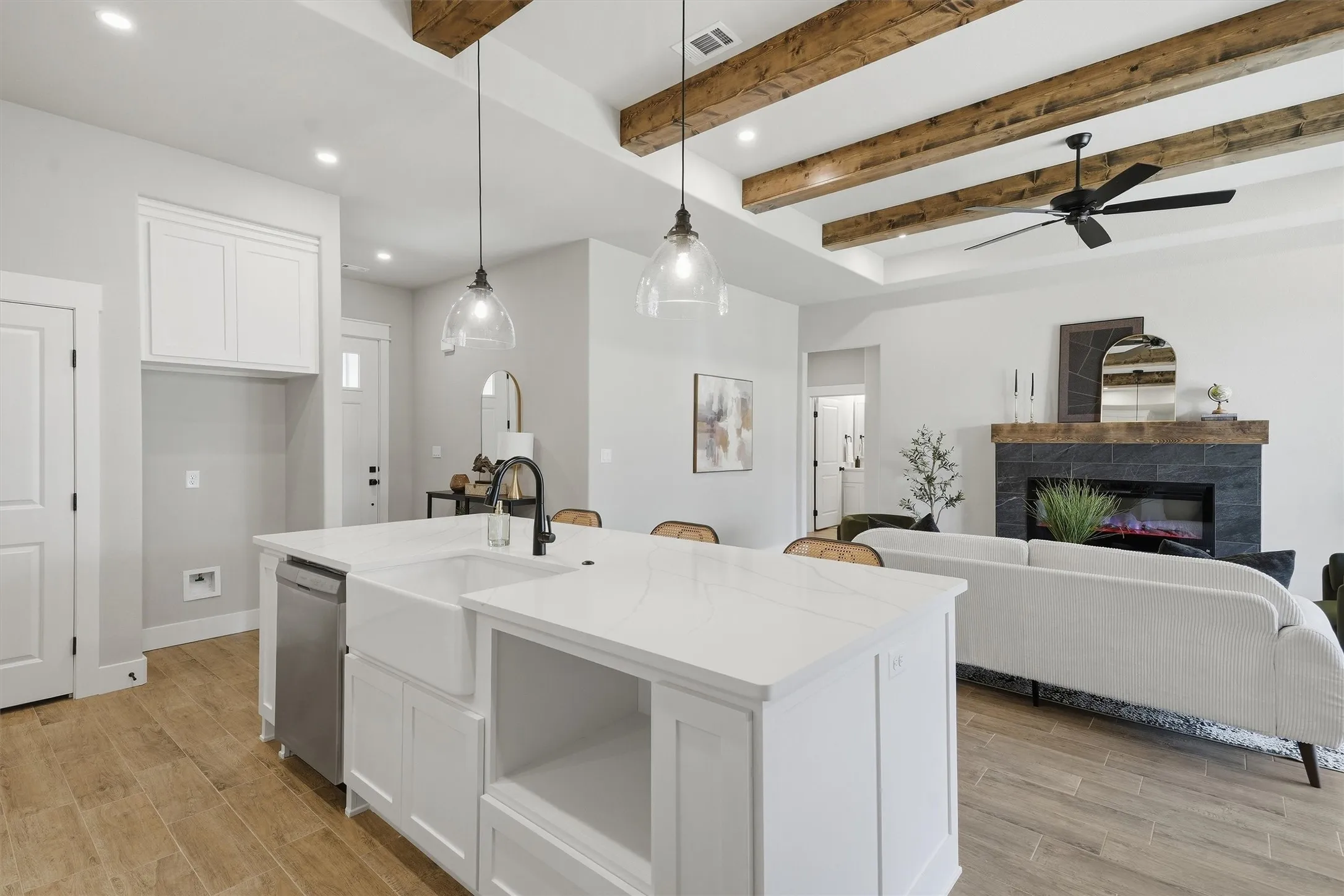 Open-concept living space with exposed wood ceiling beams, wood-finish flooring, a kitchen island featuring a farmhouse sink, and a fireplace with a dark tile surround and wood mantel