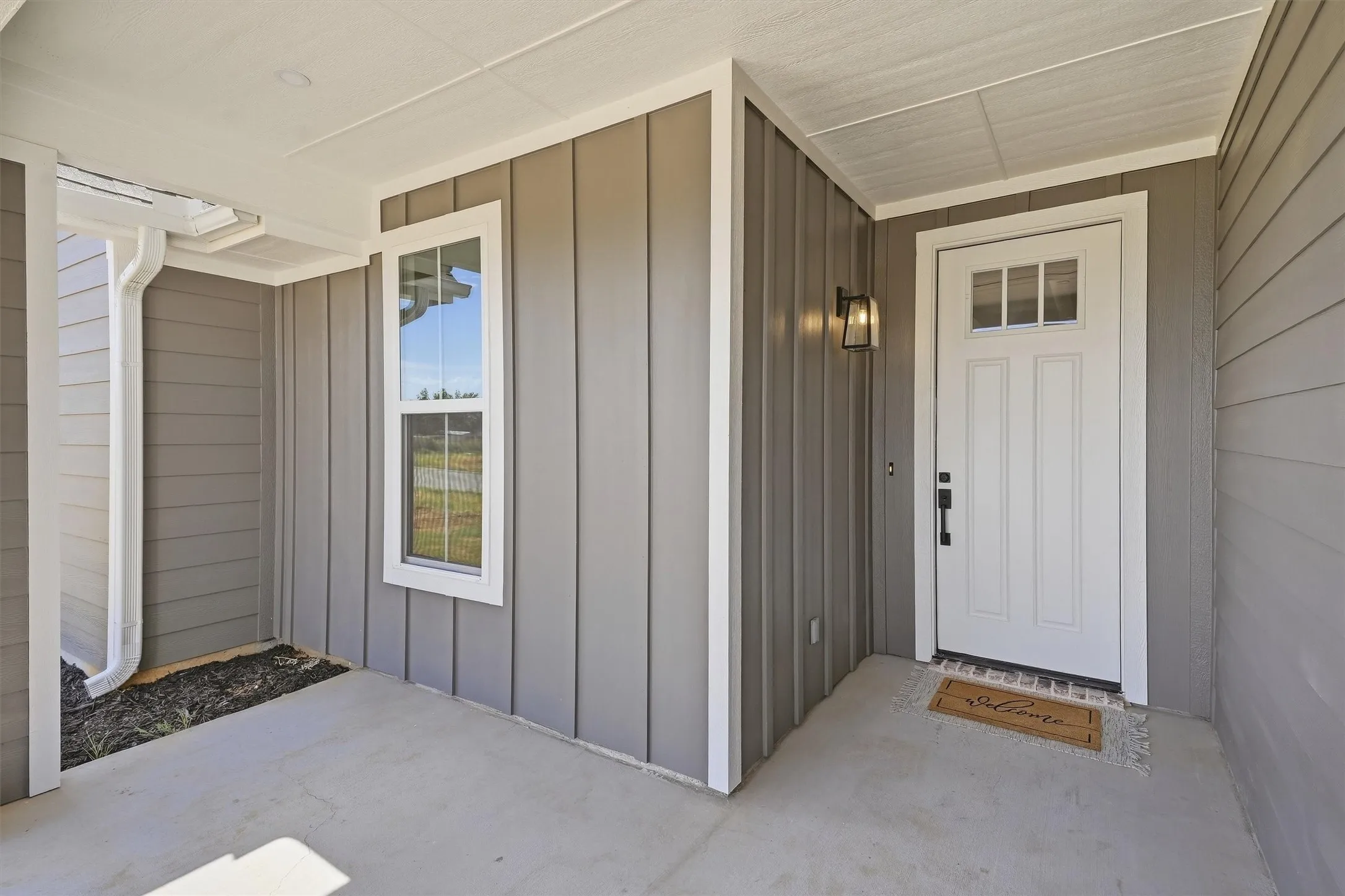 Covered front porch with concrete flooring, board and batten siding, horizontal siding, exterior lighting fixture, and a white entry door with transom windows