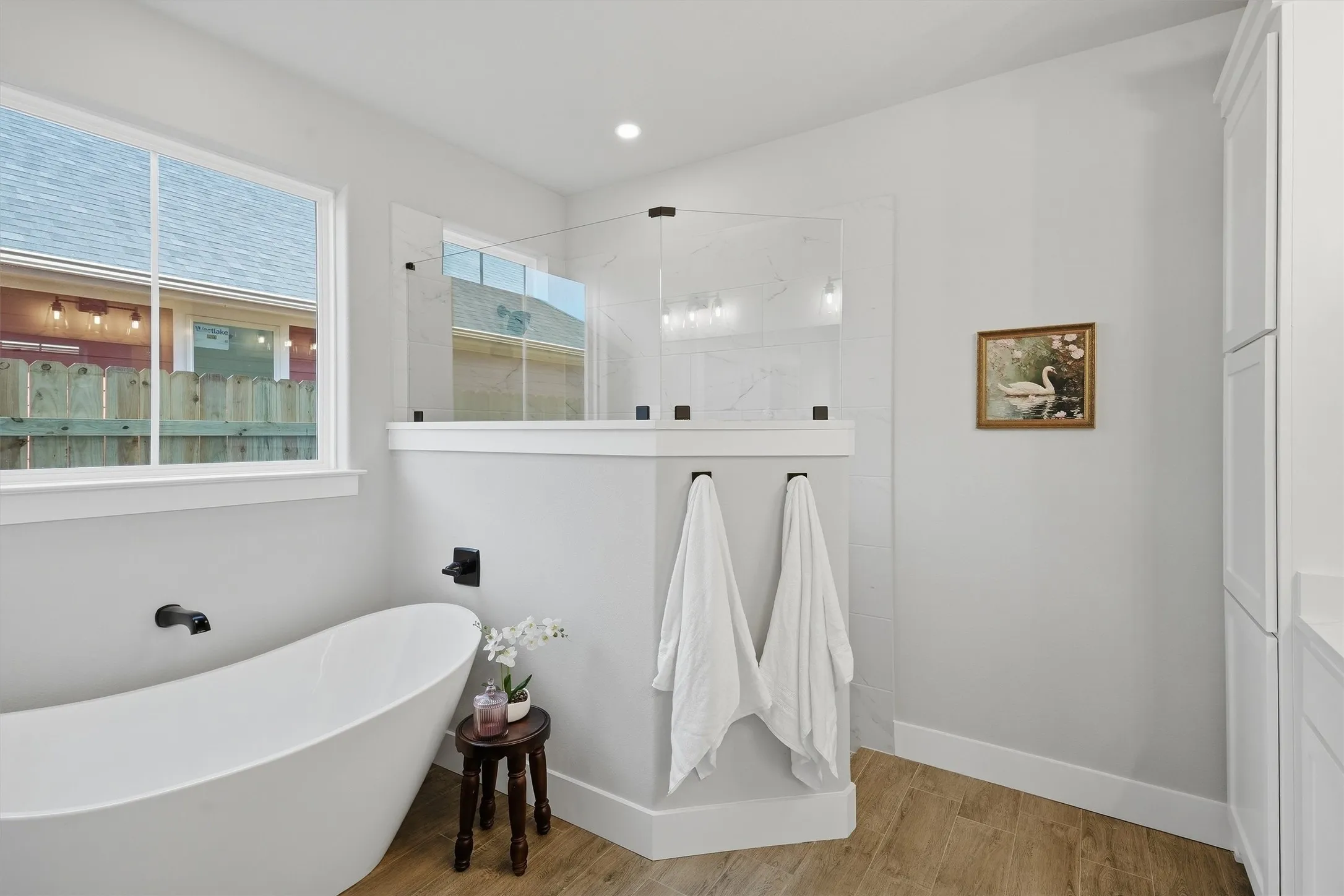 Freestanding soaking tub with matte black fixtures, glass-enclosed shower with tile surround, wood-finish flooring, and built-in white cabinetry