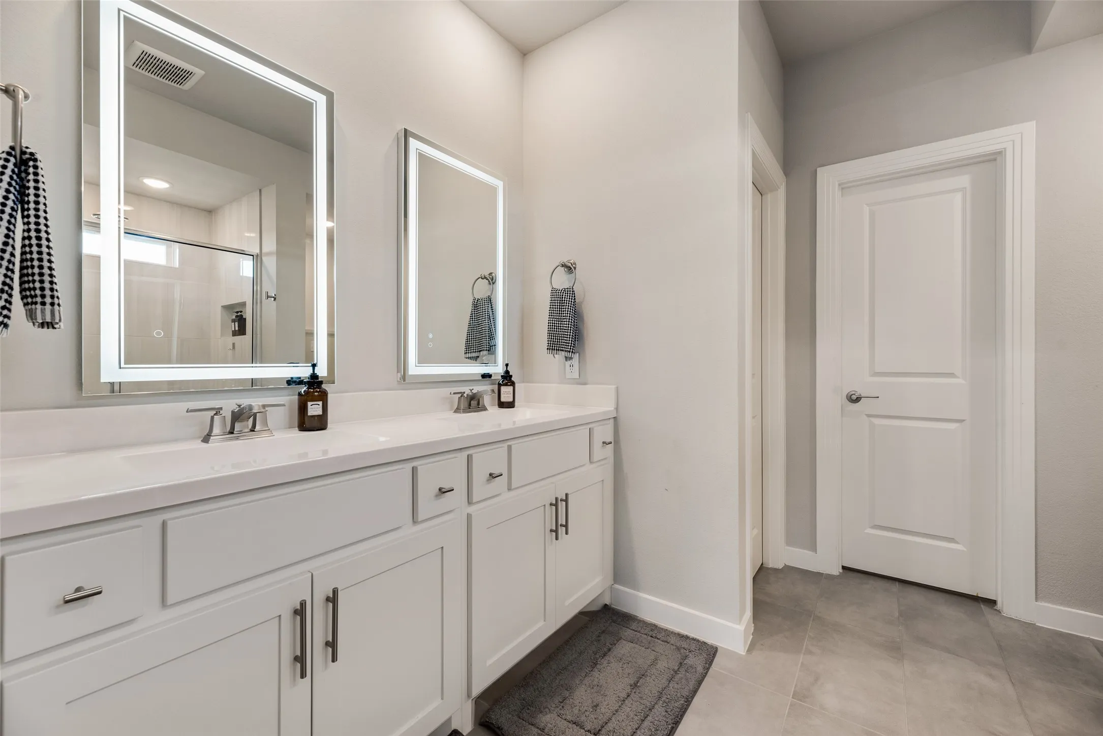 Dual illuminated vanity mirrors over a double sink, white cabinetry with silver hardware, and light-toned flooring