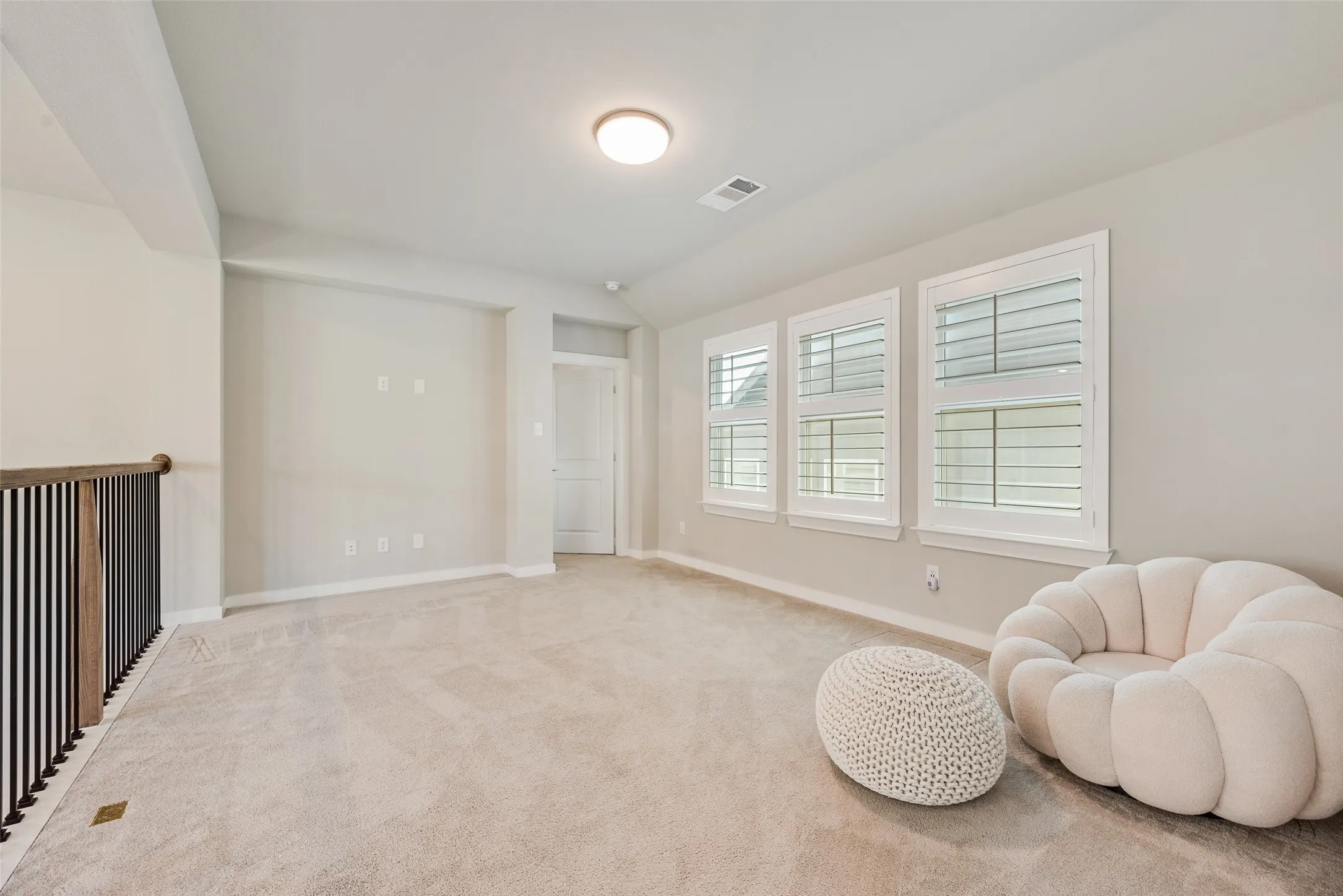 Spacious carpeted room featuring a ceiling-mounted light fixture, three windows with white plantation shutters, and a dark wood and iron railing