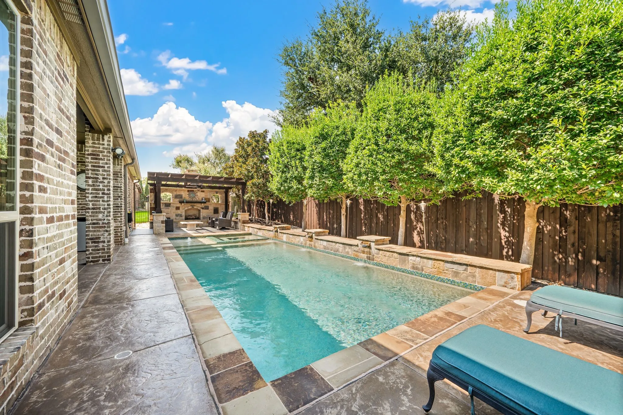 Rectangular swimming pool with stone coping, stamped concrete patio, and an outdoor living area featuring a stone-clad fireplace under a wood pergola