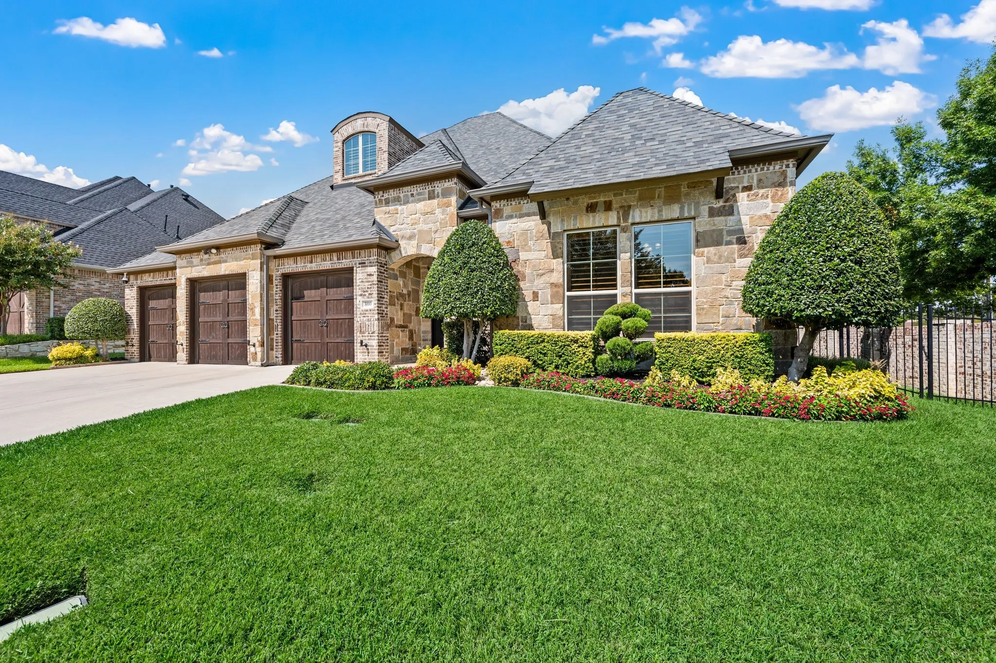 Stone facade with a prominent dormer, three wood-paneled garage doors, and a concrete driveway