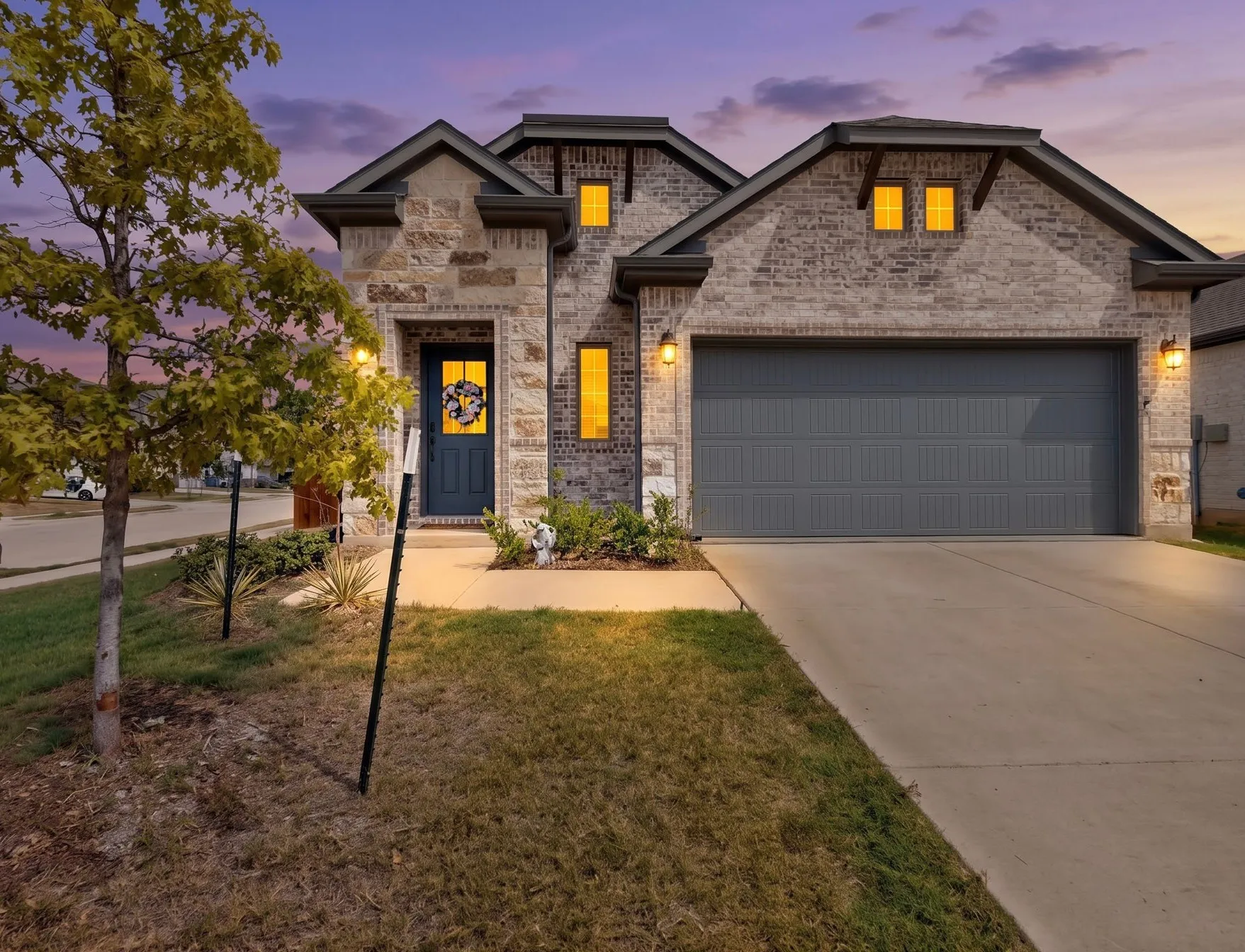 Brick and stone facade, attached garage, concrete driveway, dark entry door, and decorative exterior lighting