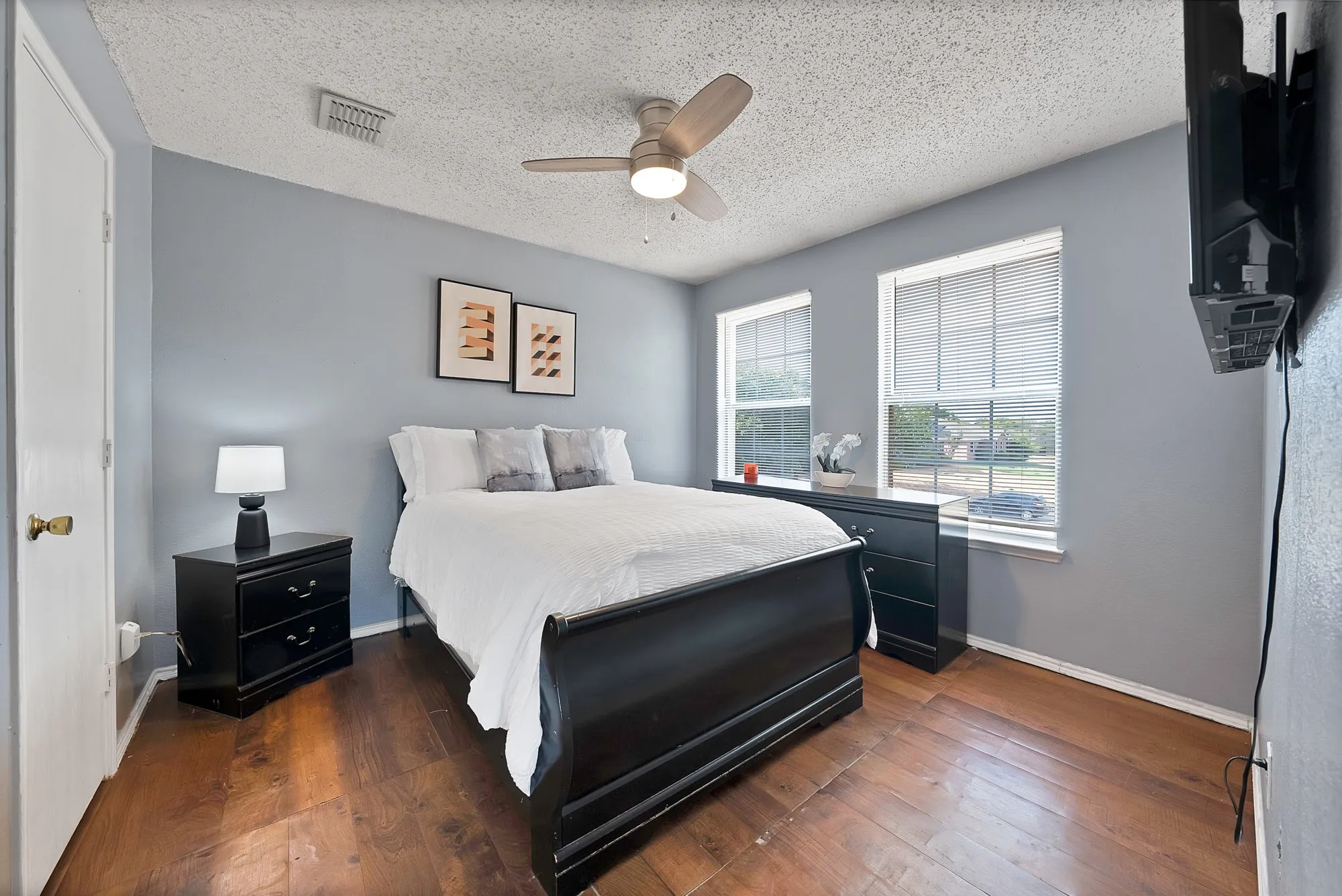 Hardwood-finish flooring, two large windows with blinds, ceiling fan with integrated lighting, textured ceiling, and light blue wall paint