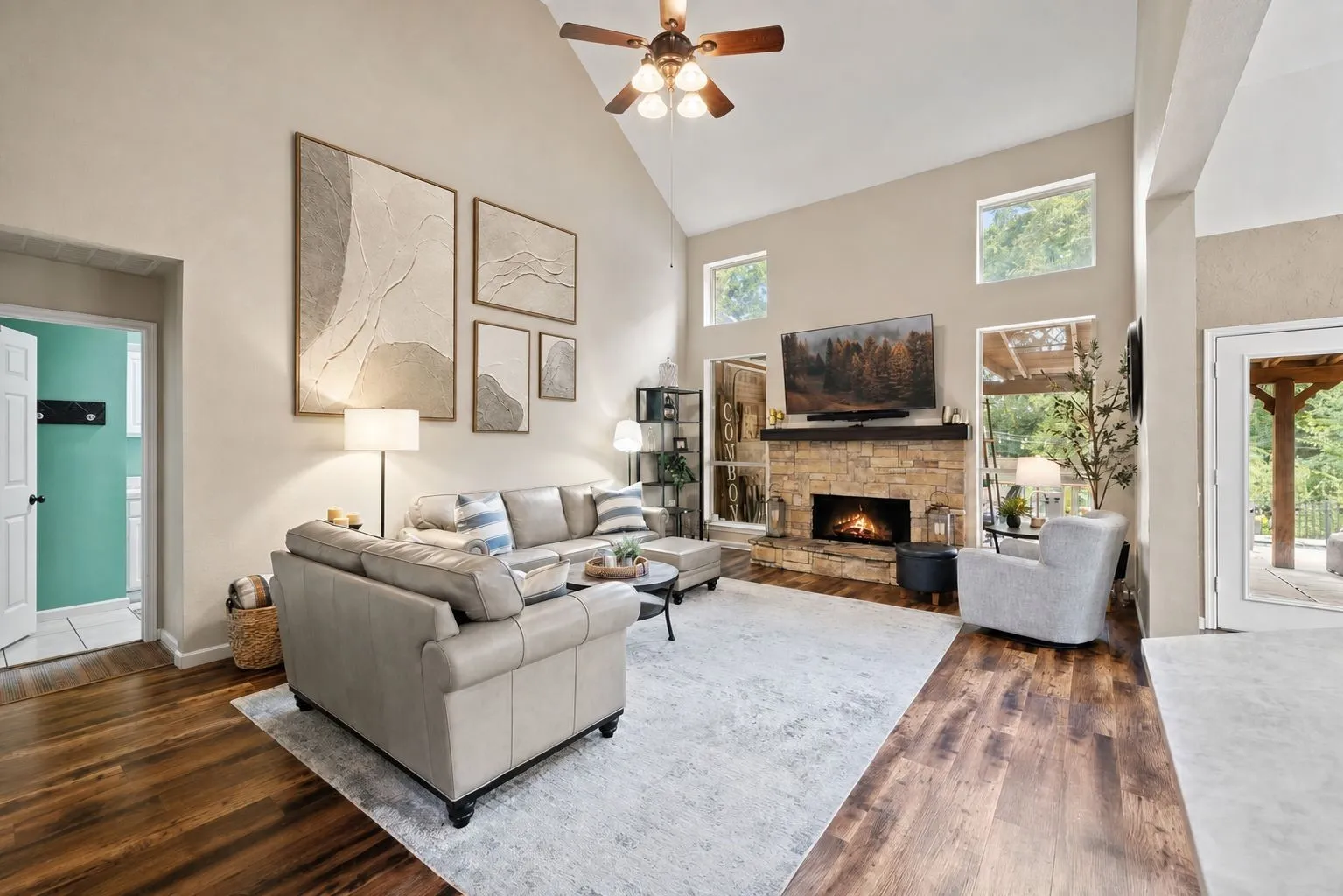 Vaulted ceiling with a ceiling fan, stone-clad fireplace with a wood mantle, wood-finish flooring, and multiple high windows