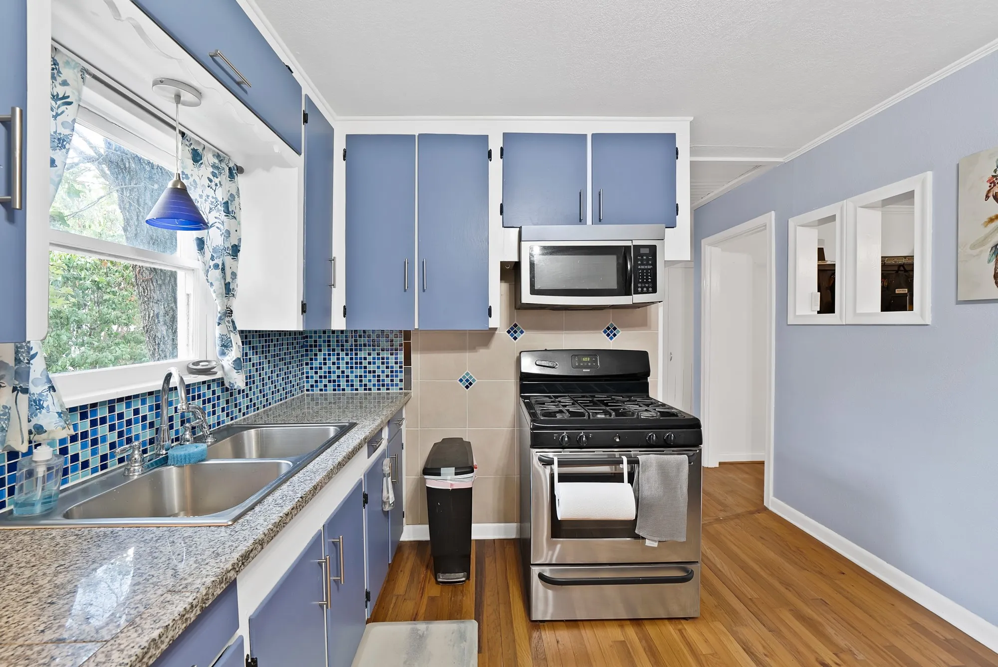 Kitchen with wood-finish flooring, blue cabinetry, mosaic tile backsplash, and stainless steel appliances including a range and microwave