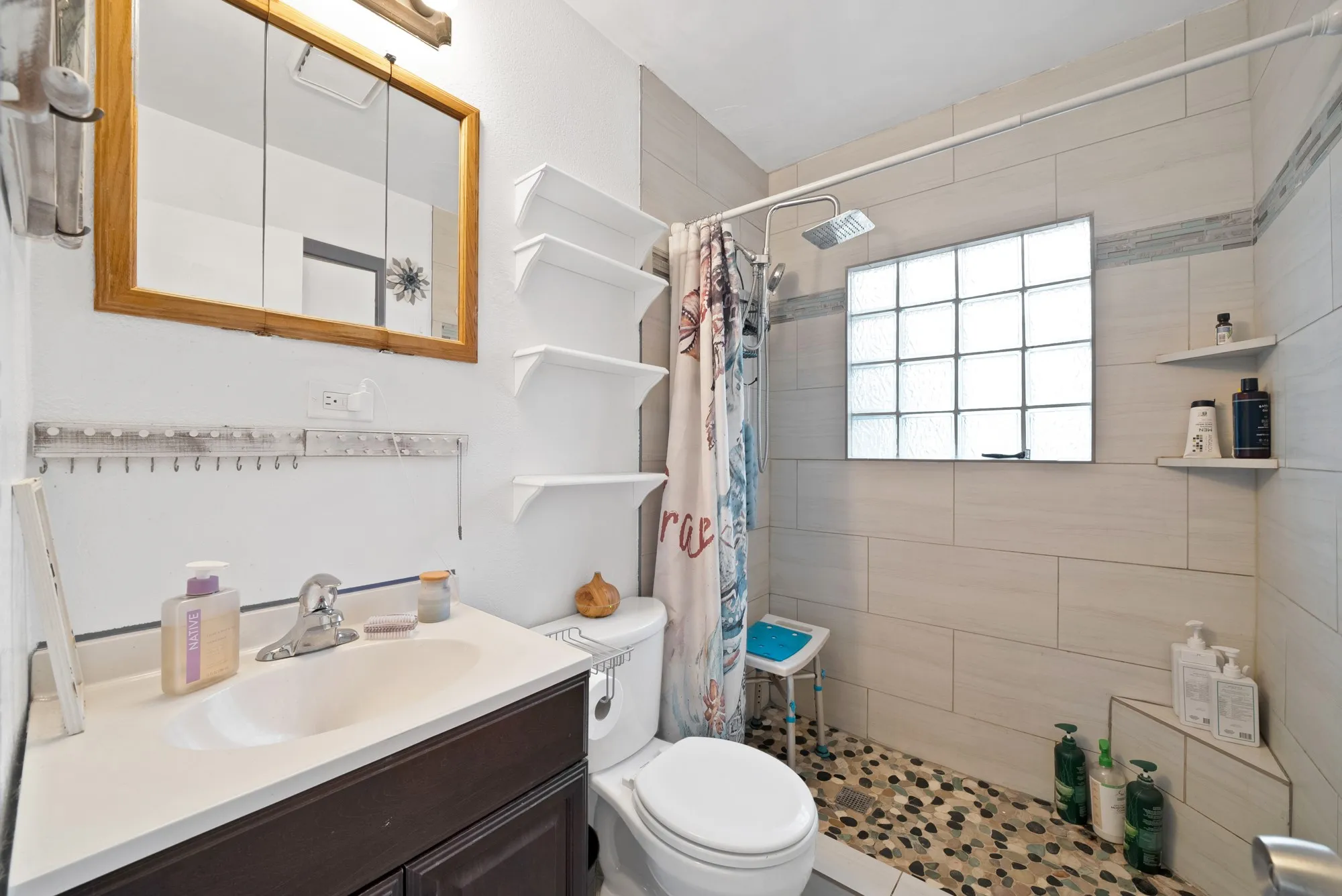 Spacious bathroom featuring a glass block window, stone pebble shower floor, tiled shower walls, and vanity with an integrated sink