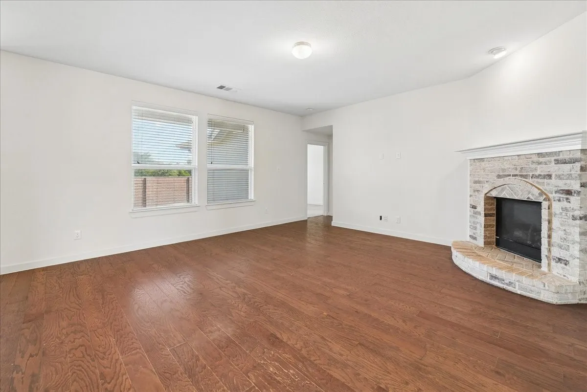 Spacious living area featuring wood-finish flooring, a brick fireplace with a hearth, and two windows with blinds