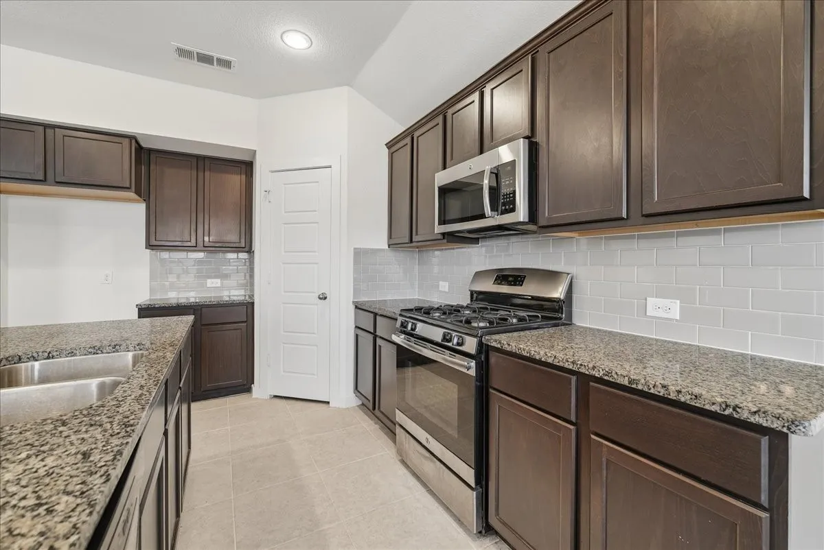 Kitchen featuring dark wood-finish cabinetry, light stone-finish countertops, stainless steel appliances, a subway tile backsplash, and a double basin sink
