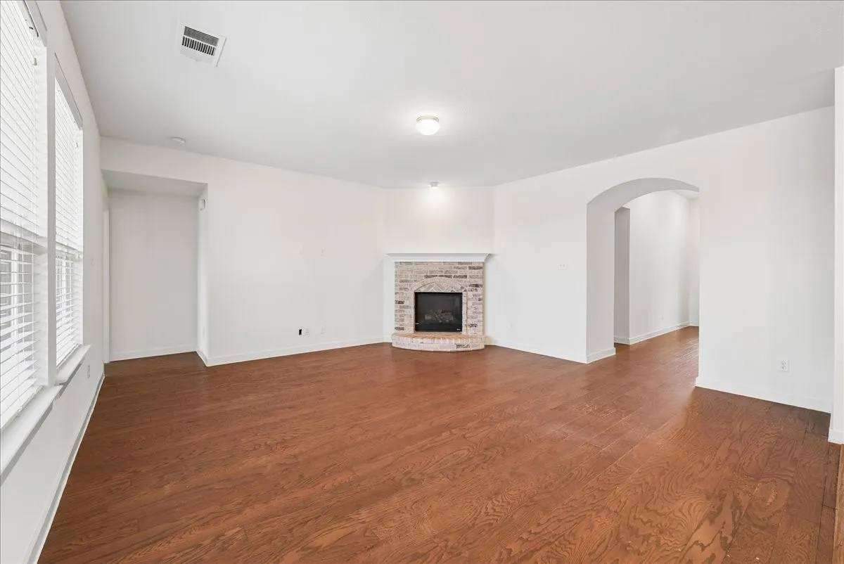 Spacious living area featuring wood-finish flooring, a corner brick fireplace, multiple windows with blinds, and an arched entryway
