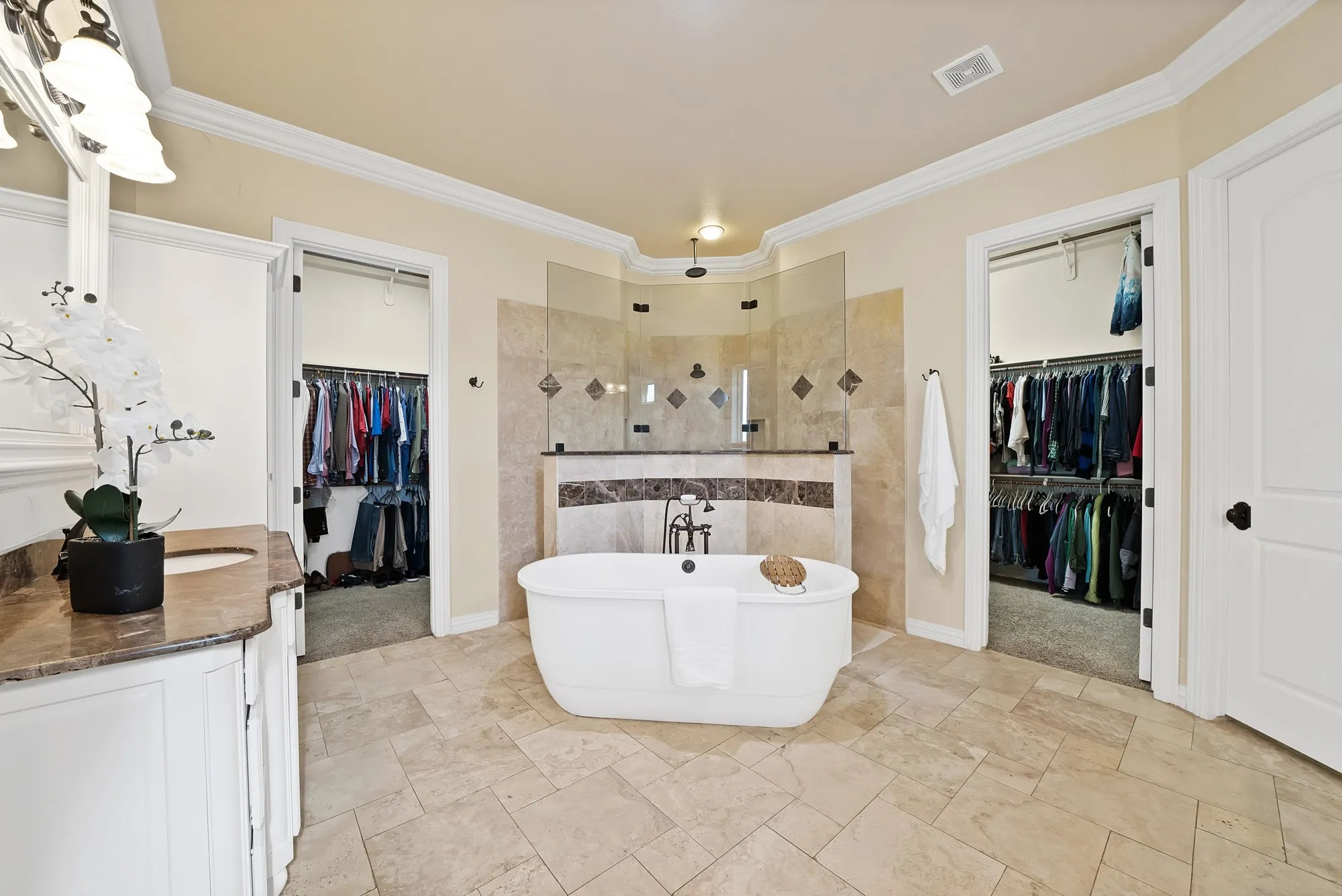 Freestanding soaking tub with dark bronze fixtures, glass enclosed shower, dual walk-in closets, single vanity with stone countertop, and crown molding