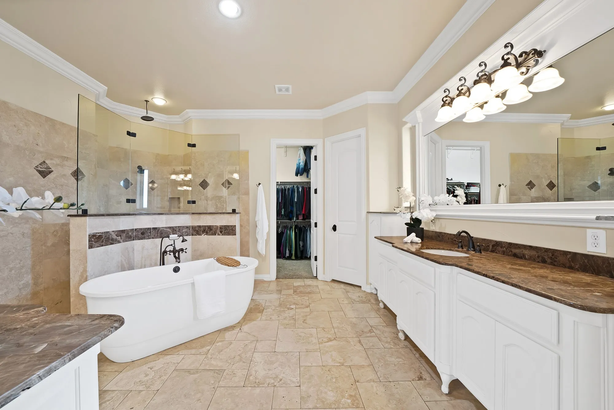 Freestanding soaking tub, large glass-enclosed shower, dual vanity with undermount sinks, stone tile flooring, and a walk-in closet